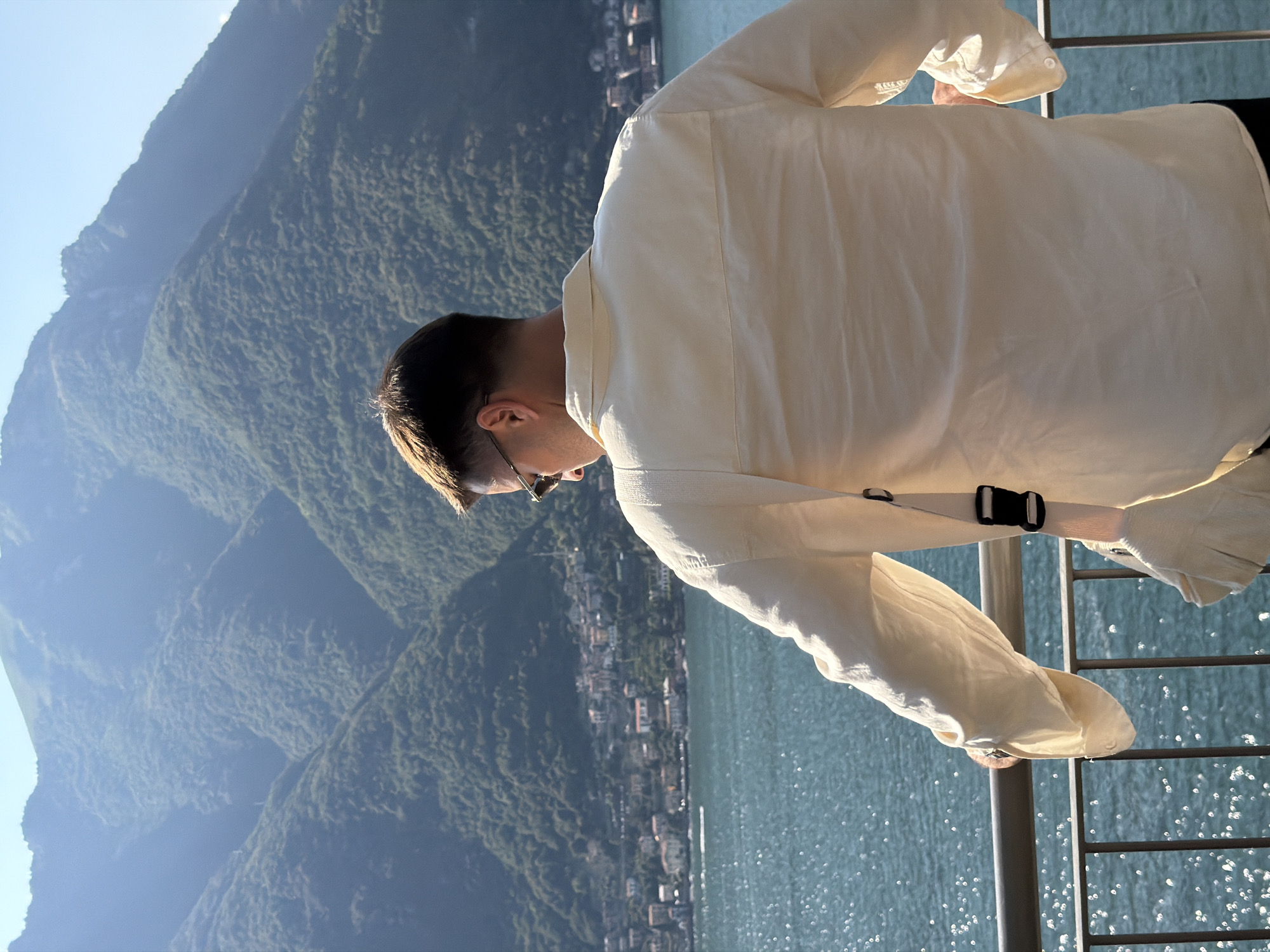 A man stands facing away from the camera, leaning on a railing, looking out at a body of water and a mountain range dotted with buildings. The man is wearing a light-colored, long-sleeved collared shirt and sunglasses. A strap from a bag is visible across his back and shoulder. The water is a bright blue with sunlight reflecting off its surface. The mountains in the background are covered in green vegetation, with a town or village nestled along the shoreline. The sky is clear and blue. The scene appears to be a scenic overlook in Pognana Lario, Italy.