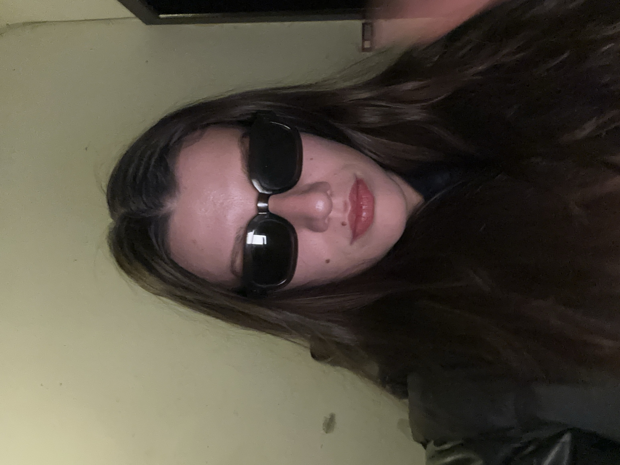 A woman with long, wavy brown hair and fair skin is facing the camera while wearing black sunglasses. She has visible moles on her face and is wearing lipstick. The background appears to be a wall of light yellowish-green color and an undefined object in the upper part of the image. The scene provides no information regarding the city of Burgas, Bulgaria.