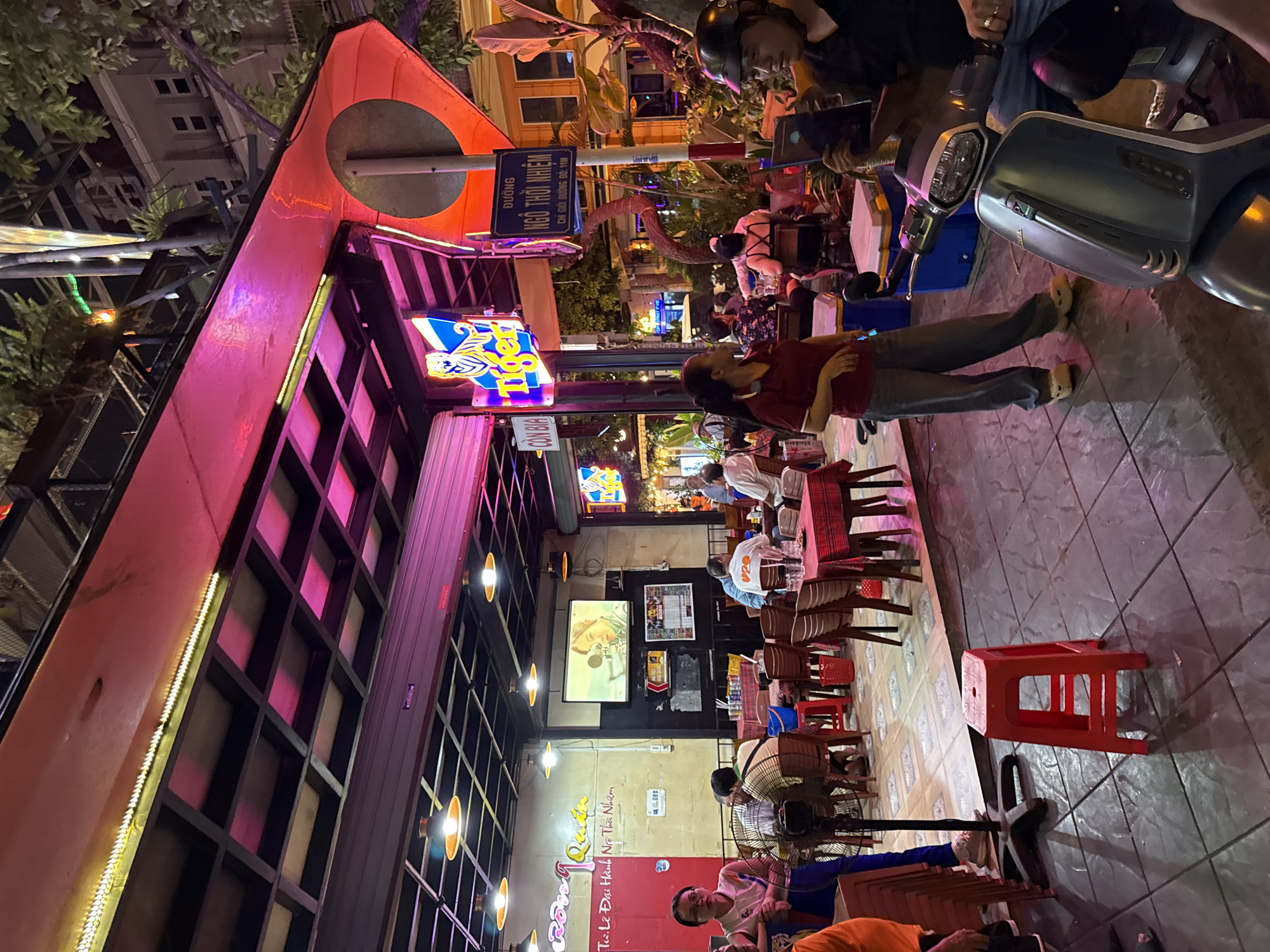 The image depicts a nighttime scene at an outdoor establishment in Nha Trang, Vietnam. The establishment appears to be a restaurant or bar, with tables and chairs set up on a tiled patio. A red plastic stool is prominently placed in the foreground. The area is lit by overhead lamps and neon signage, including a "Tiger" beer sign and another sign with Vietnamese text. In the background, people are seated at tables, some eating and drinking. A television screen displays a person. A scooter is parked on the right side of the image, with a person seated on it, holding a tablet. The overall atmosphere is lively and casual, typical of a street-side eatery in a Vietnamese city.