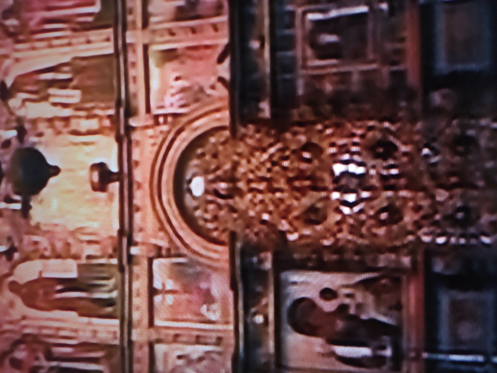 The image shows a section of an ornate ceiling. It features a grid-like structure with framed panels containing faded paintings or frescoes of human figures. There is a large circular decorative element with an intricate, possibly mosaic-like, design at the center. The surrounding structure appears to be made of dark wood or metal with rivets or decorative elements. The lighting is dim, and the overall color palette is muted with shades of brown, gold, and faded reds. No information in the image suggests the location is in the city of Pavlodar, Kazakhstan.
