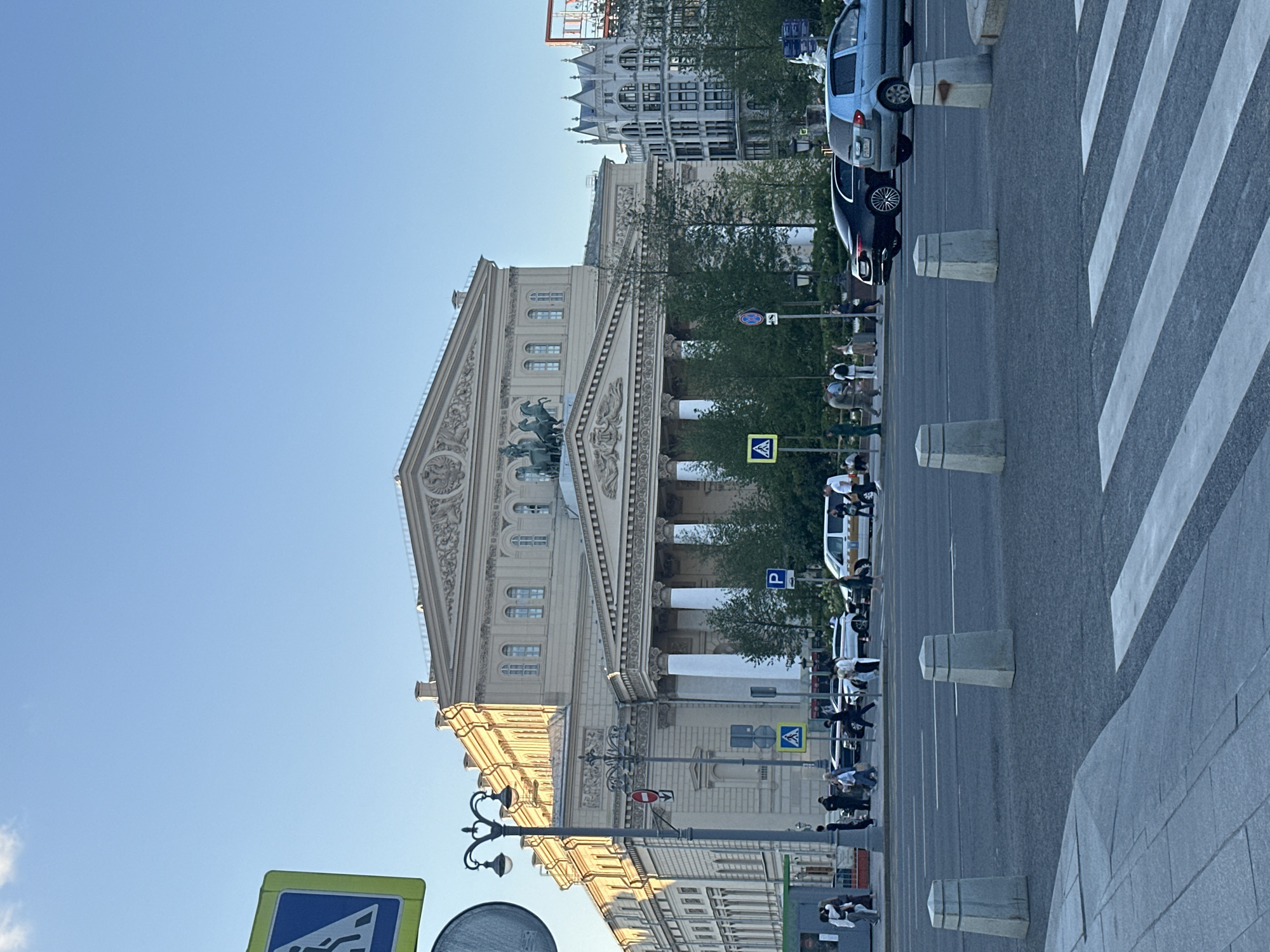 This is a photograph of the Bolshoi Theatre in Moscow, Russia, taken from across a street. The imposing neoclassical building dominates the background, its grand facade featuring a pediment topped with a sculpture of Apollo. Rows of tall columns support the portico. The building is bathed in the warm light of what appears to be late afternoon or early evening.
In the foreground, a street with a pedestrian crossing is visible. Several cars, including a black sedan and a light blue hatchback, are on the road. Pedestrians are walking on the sidewalk and crossing the street. Streetlights, traffic signs, and concrete bollards are also present. The sky is clear blue with a few wispy clouds. To the right of the Bolshoi Theatre, a more modern-looking building with Gothic architectural elements can be seen. The overall scene depicts a busy urban environment with a historic landmark as its centerpiece.