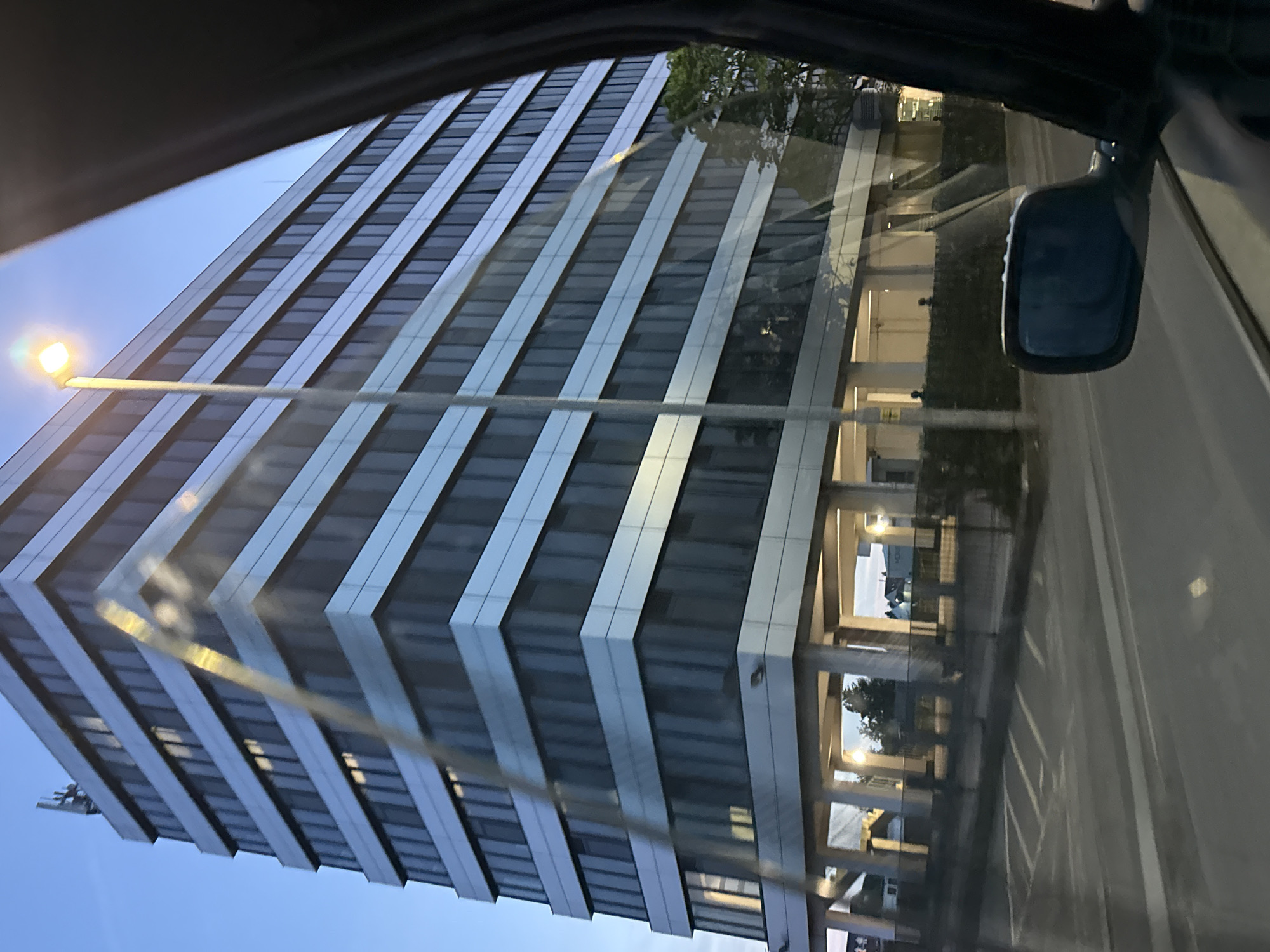 The image depicts a view from inside a vehicle, likely a car, looking out at a building in Great Yarmouth, United Kingdom. The building is a tall, modern structure with multiple floors, characterized by alternating horizontal bands of dark glass and light-colored paneling. A street lamp is visible in the foreground near the building. The building's reflection is somewhat visible on the car's windshield, obscuring a parking area and a walkway lined with columns. The car's side mirror is also visible. The sky is visible, transitioning from a light blue towards the horizon. The overall setting suggests an urban or suburban environment. The image does not provide sufficient detail to identify a specific location within Great Yarmouth.