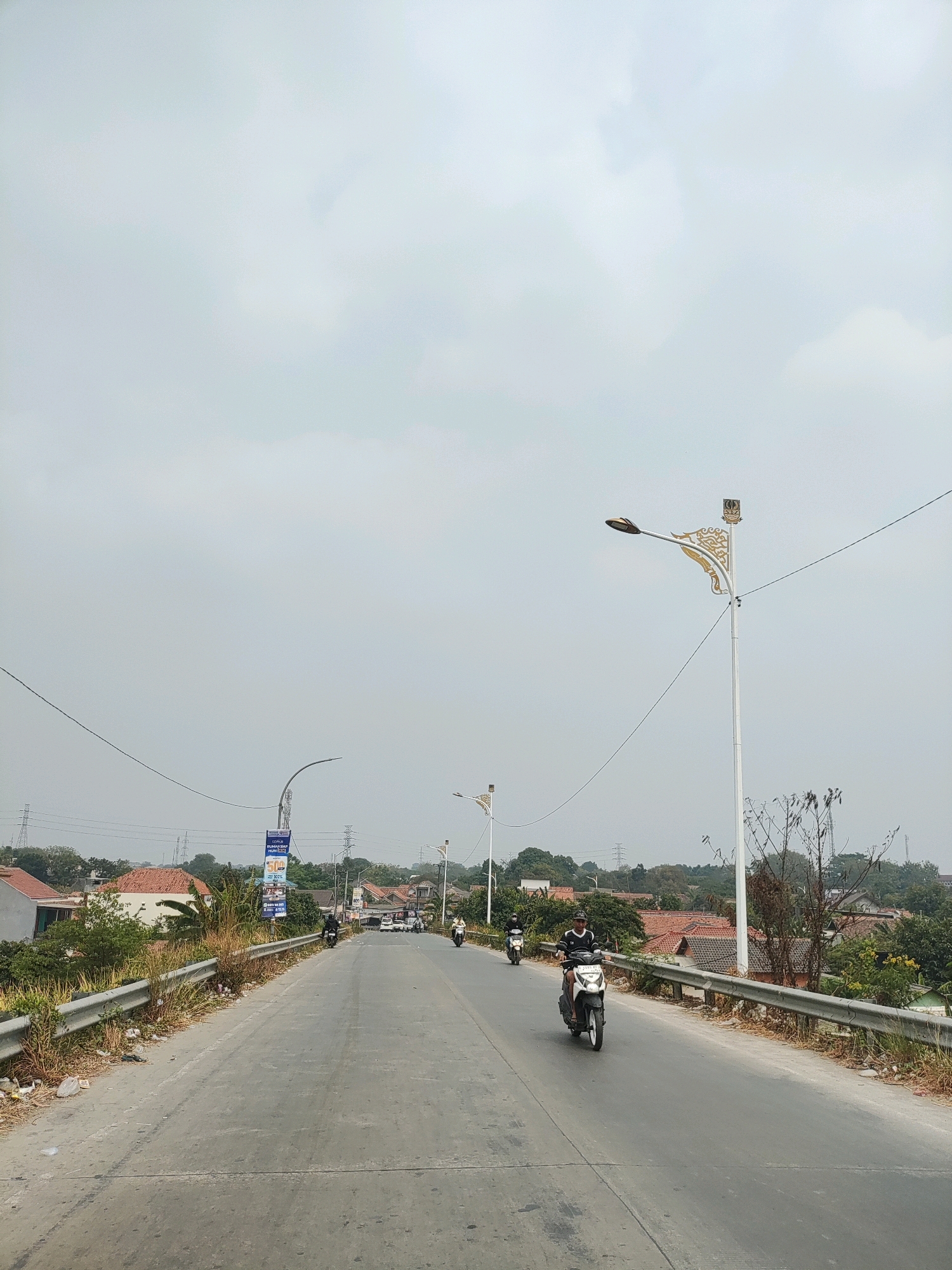 The image depicts a view of a road in Wanajaya, Indonesia, under an overcast sky. The road is paved and appears to be a two-lane street with a median marking. On the right side of the road, there is a metal guardrail, and beyond it, a residential area with houses featuring red tile roofs and lush green trees. Several power lines stretch across the scene, supported by utility poles.
In the foreground, a person is riding a white scooter, wearing a dark and white striped shirt and a black helmet. Further down the road, another person is riding a scooter, also wearing dark clothing. In the distance, more scooters and some cars are visible on the road, indicating ongoing traffic. Two decorative streetlights with intricate golden designs are prominently featured along the road. One streetlight is in the mid-ground on the right, and another is partially visible further down on the left. A billboard with text and images is also present near the left side of the road. The overall environment suggests a suburban or semi-urban setting in Indonesia.