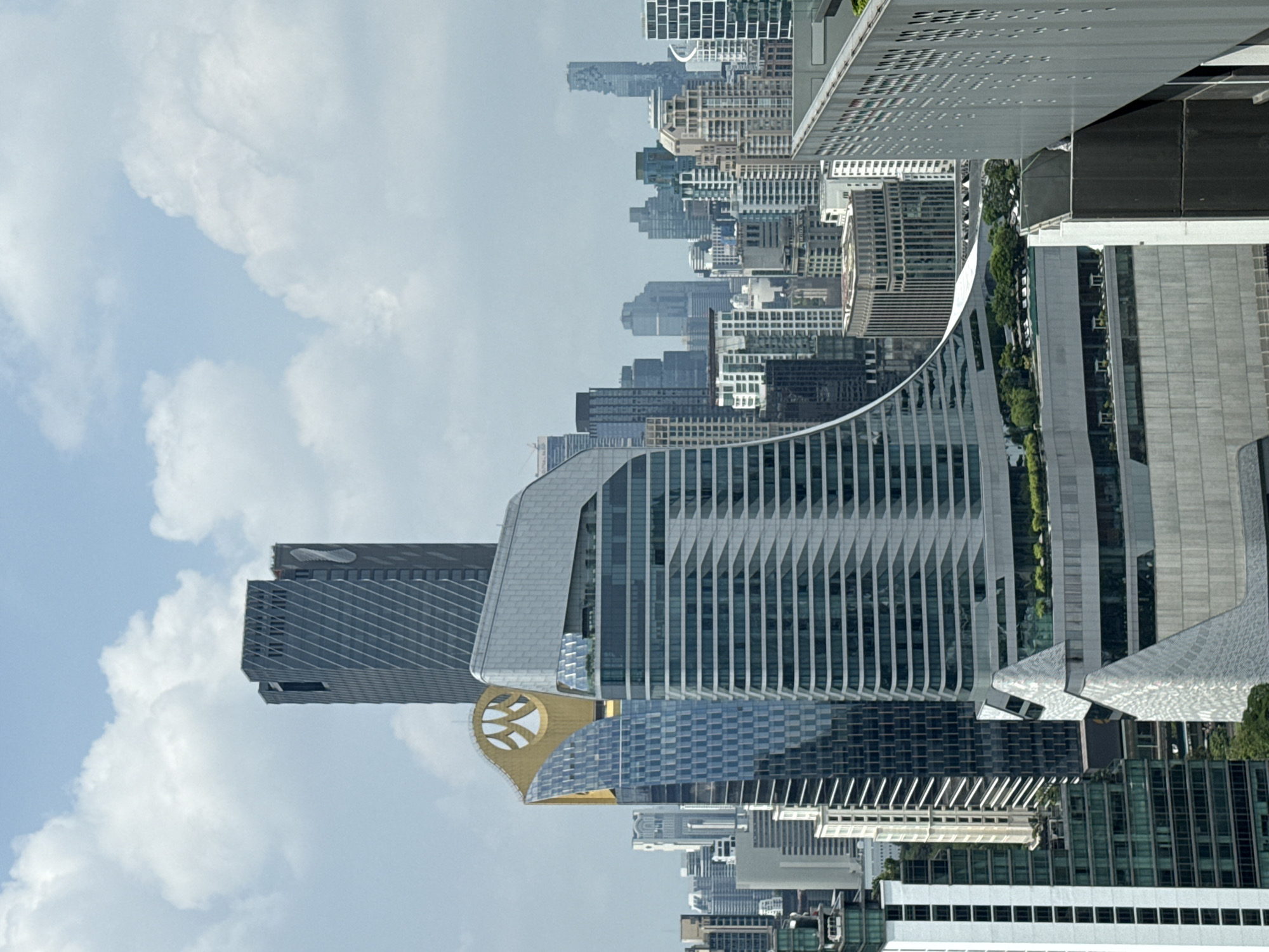The image shows a daytime view of the Bangkok skyline in Thailand, taken from an elevated position. Prominent buildings include the MahaNakhon tower, the curved structure of the Rosewood Bangkok hotel, and various other high-rise buildings. The sky is partly cloudy. The photograph is rotated 90 degrees counterclockwise.