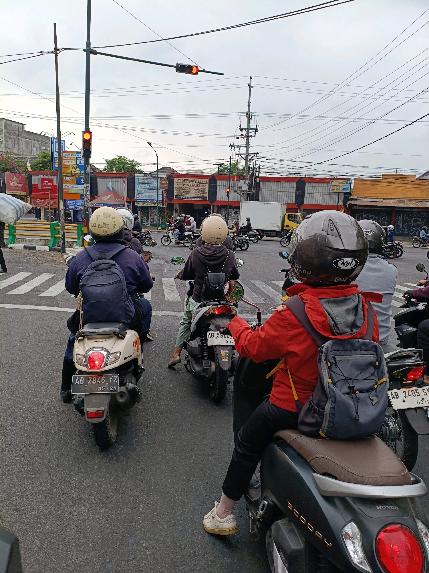 This is a medium shot of a traffic intersection in Yogyakarta, Indonesia, on a cloudy day. The perspective is from behind several motorcyclists stopped at a red light. In the foreground, a person wearing a red jacket and a dark backpack sits on a dark colored scooter, their back to the camera. To the left, another motorcyclist in a blue jacket and a dark backpack is stopped on a cream-colored scooter. To the right and behind, another person in a grey shirt is on a scooter. In the background, various buildings with signage line the street, including a yellow truck, a white truck, and several other motorcyclists. Traffic lights, power lines, and utility poles are visible overhead. The road is asphalt with white pedestrian crossing lines. License plates are visible on the scooters, with one reading "AB 2846 YZ".