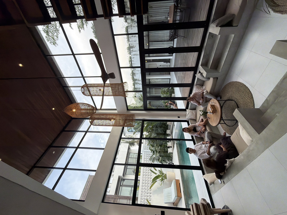 The image displays a modern, high-ceilinged living space within a residence located in the city of Unknown, Indonesia. The indoor area features a U-shaped, built-in concrete sunken lounge with light beige cushions and pillows. Five individuals are seated on the lounge. Two men and three women are present, dressed in casual clothing. They are positioned around a round wooden coffee table, which holds a small wooden box, a clear glass, and a small vase containing white flowers, all resting on a round woven rug.
The high ceiling is paneled with dark wood, from which three large, woven natural fiber pendant lights and one large, dark wooden ceiling fan are suspended. The living space is bordered by floor-to-ceiling black-framed glass panels and sliding doors, offering a view of an outdoor area.
Outside, a rectangular swimming pool with blue water is directly adjacent to the glass. A light-colored lounge chair with a cushion is positioned by the pool. Tropical plants are arranged in planters and beds along a textured white wall. Beyond the pool, a dark wooden deck extends, featuring an outdoor dining set comprising a table and dark woven chairs. One individual is partially visible outdoors, seated on the edge of the pool deck or steps. White tiled flooring covers the non-sunken indoor areas, and a dark wooden open-tread staircase is visible on the far right. The scene is illuminated by natural daylight.