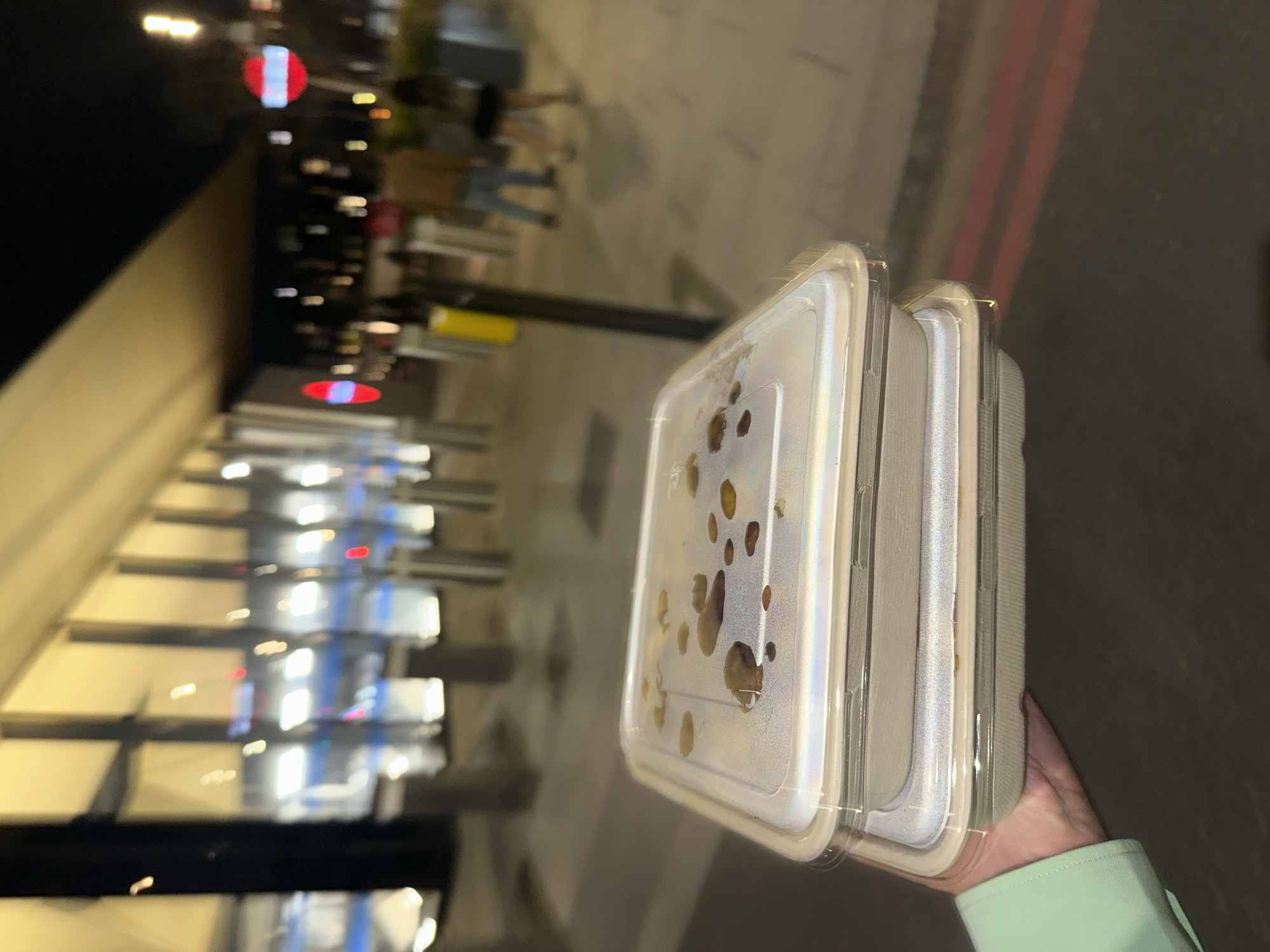 The image shows a hand holding three takeaway containers in an outdoor setting. The hand is wearing a pale green sleeve. The containers appear to have clear lids with condensation and some contents visible inside. The background features blurred lights, suggesting an urban environment. There are reflections of lights on what looks like a building façade with repetitive architectural elements. Based on the visual cues of architecture, street lighting, and the presence of takeaway food containers, this scene could be in London, United Kingdom. There are indistinct figures in the background, suggesting people are present. The scene is illuminated, possibly at night. There is a road or sidewalk nearby, defined by a red line. The primary action is the holding of the takeaway containers, implying someone has recently obtained or is transporting food.