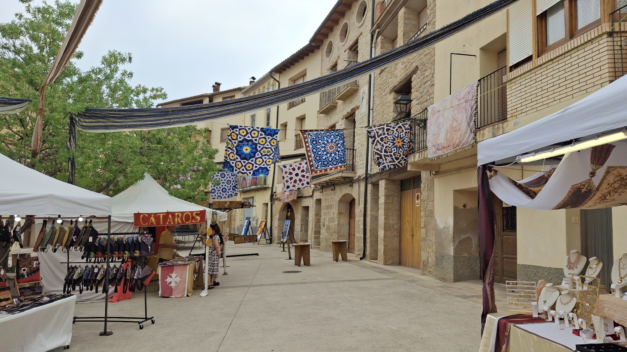 This image depicts a street scene in La Fresneda, Spain, likely during a market or festival. The foreground features market stalls with white tents. One stall displays various bags and leather goods hanging from racks. Another stall, marked with a red sign that reads "CATAROS," exhibits a display of jewelry on mannequins and stands. A woman in a patterned dress is seen interacting with the merchandise at one of the stalls. In the background, buildings with stone facades and balconies line the street. Colorful, intricately patterned banners or tapestries are hung across the street and from the balconies, adding to the festive atmosphere. Trees with green foliage are visible, and the sky is overcast. The ground is paved.