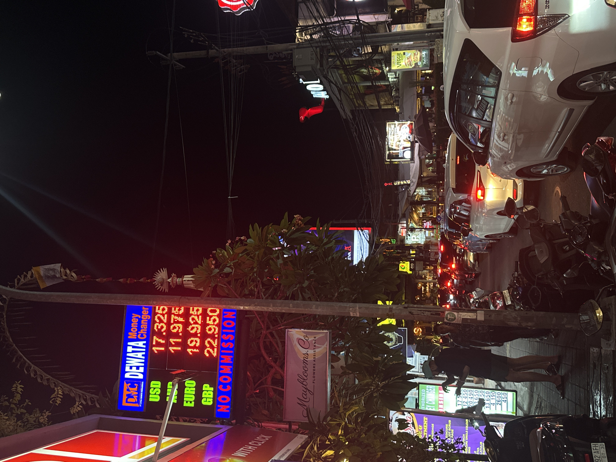 The image depicts a busy street scene at night in Kuta, Indonesia, likely a tourist area. A prominent money changer sign displays exchange rates for USD, AUD, EURO, and GBP, with the text "NO COMMISSION" in large, bright blue letters. To the left of the money changer, a sign for "Mayblooms. Co FLOWERS ASSISTANCE" is visible. The street is congested with traffic, primarily white cars and motorcycles. Several individuals are present, including a man in a teal cap and dark clothing standing on the sidewalk and gesturing towards a sign. Another man in a patterned shirt stands nearby. The environment is characterized by streetlights, neon signs, and the dark night sky, suggesting an urban setting with commercial activity. Overhead, electrical wires crisscross the sky, indicating a developed infrastructure. The overall atmosphere is lively and active, typical of a commercial district at night.