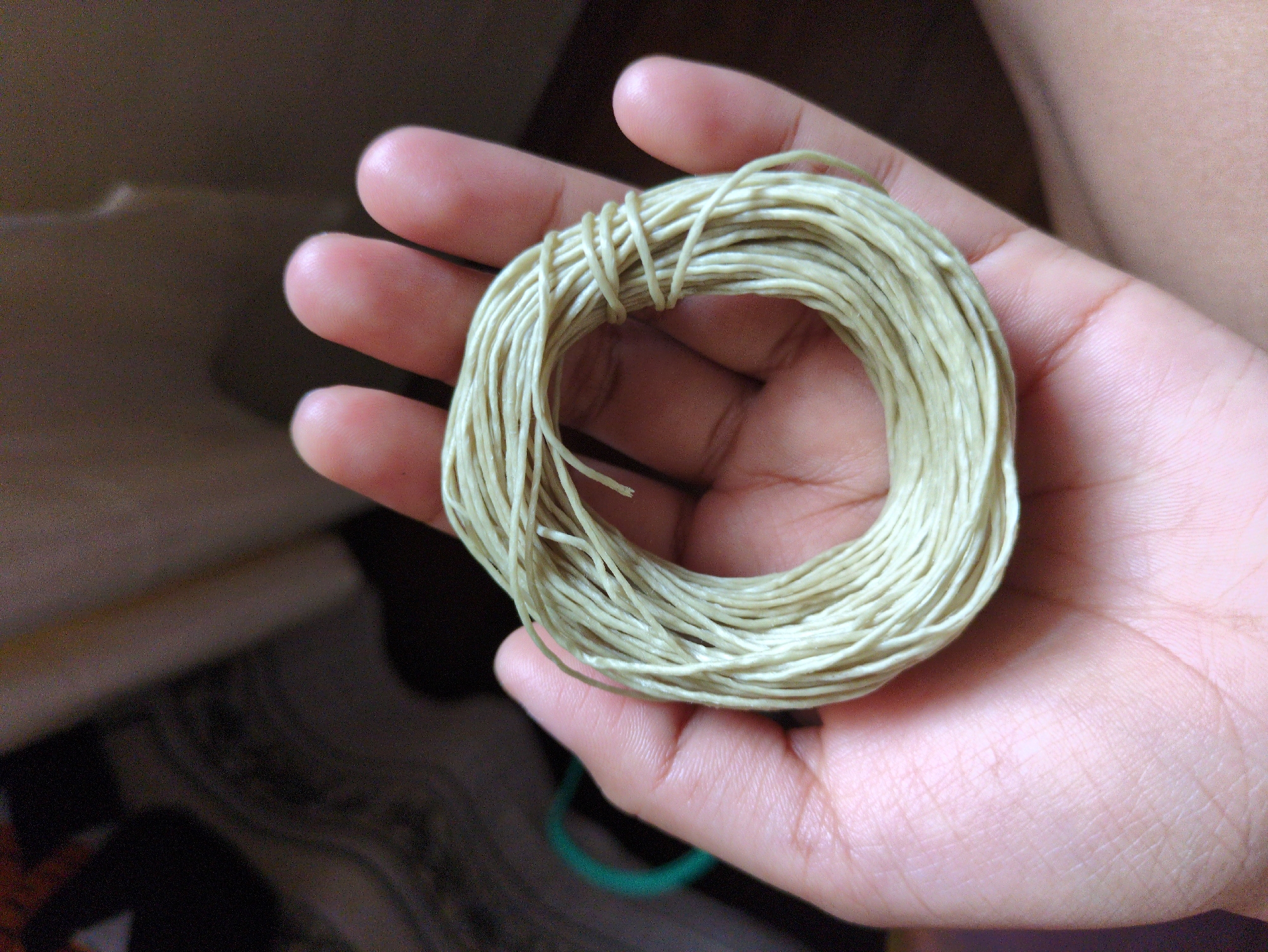 The image shows a close-up view of a person's hand holding a coil of light green string. The string is thin and appears to be made of a natural fiber, possibly jute or cotton. It is wound into a compact, circular shape, with several strands wrapped around the core. The hand is positioned to cradle the coil, with the fingers gently curved around the edges. The background is blurred and out of focus, suggesting an indoor setting with dim lighting. The wall in the background appears to be a neutral color, possibly beige or light brown. There is a hint of another object to the left of the hand, a curved, dark green item, but it is too out of focus to be clearly identified. The scene is devoid of any context that would place it in Haini, India, or any specific city.