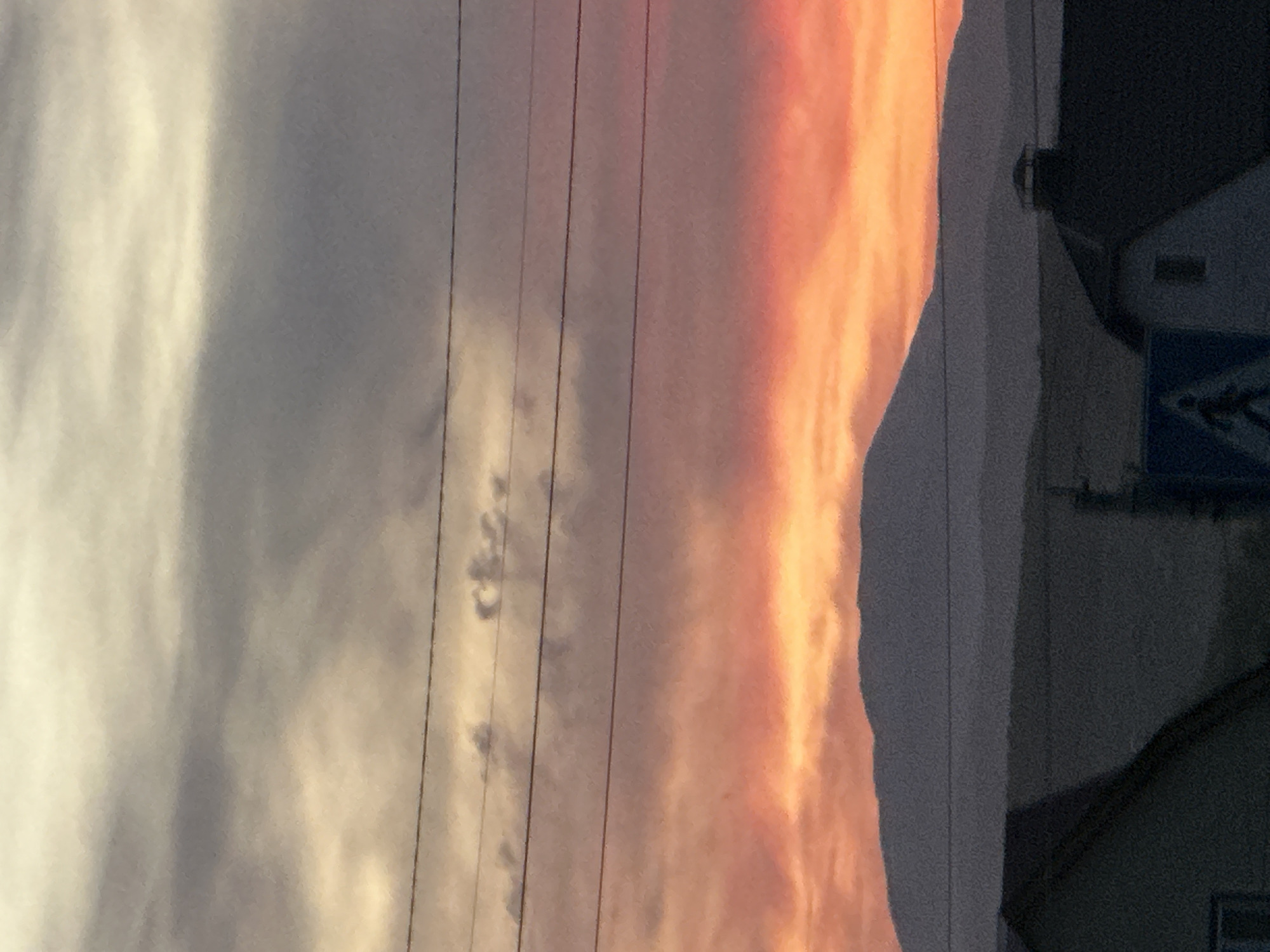 The image depicts a dusk or dawn scene in an urban area, with power lines running across the frame. To the left, the sky transitions from bright white to gray. To the right, the sky exhibits orange and pink hues, indicating either sunrise or sunset. Adjacent to the sky on the right is a dark, silhouetted structure, potentially a building or hill. Near the structure, a partially visible pedestrian crossing sign can be seen. The location is specified as a city in Unknown, Unknown.