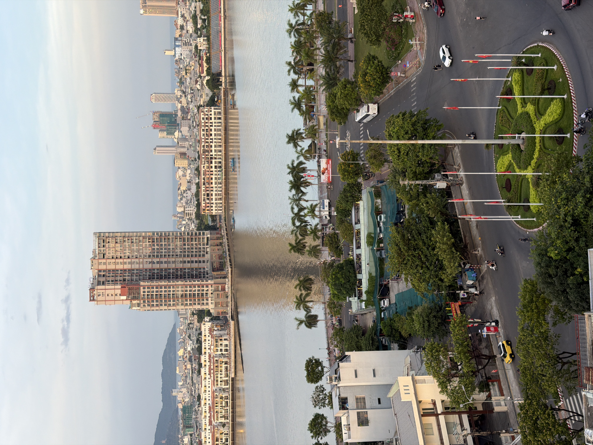 This is an aerial shot of a city scene in Da Nang, Vietnam, captured during the day. The image showcases a wide river in the foreground, with a dense urban landscape on the far side featuring numerous buildings, including several high-rise structures and smaller residential blocks. In the middle ground, a waterfront promenade is visible, lined with palm trees and smaller buildings. The foreground is dominated by a multi-lane road with a circular roundabout adorned with flags and manicured greenery. Several vehicles, including cars, motorcycles, and a bus, are navigating the roads. A few pedestrians are also visible, along with trees and some construction sites. The sky is partly cloudy, indicating fair weather. The presence of distinct architectural styles and the overall urban layout strongly suggest this is a waterfront area in Da Nang, likely near the Han River.