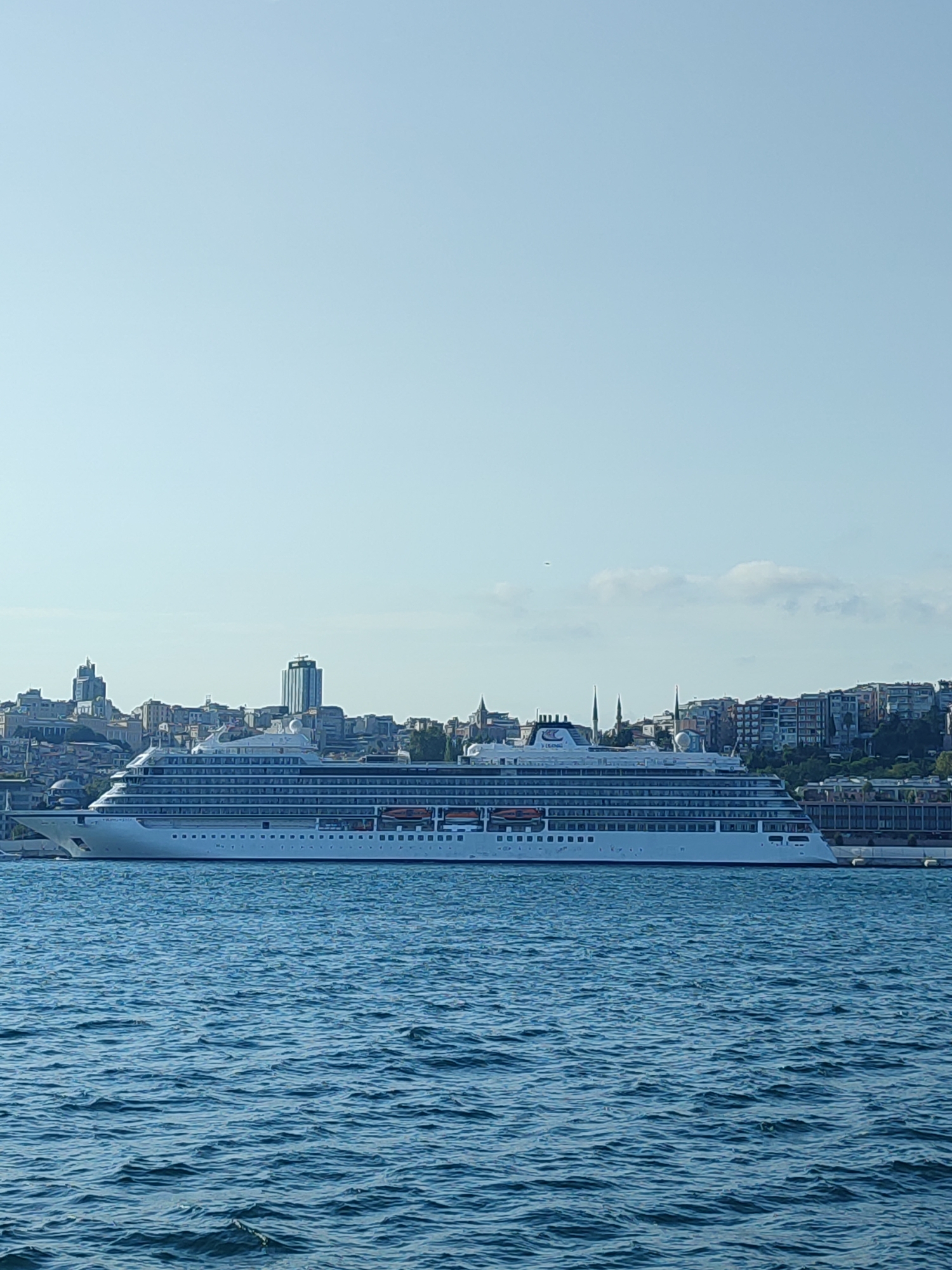 A wide, low-angle shot captures a large, white cruise ship docked in the foreground, with the city of Istanbul, Turkey, visible in the background under a clear, pale blue sky. The cruise ship, identified by the Viking logo on its side, spans horizontally across the mid-section of the image, its numerous decks and windows clearly visible. Lifeboats are positioned along the ship's upper levels.
The cityscape behind the ship features a dense collection of buildings of varying heights, including modern skyscrapers and older structures with domed roofs and minarets. Green trees dot the landscape between the buildings, suggesting a city with significant vegetation. The water in the foreground is a deep blue, with small, choppy waves creating a textured surface. The overall impression is one of a sunny day in a bustling port city.