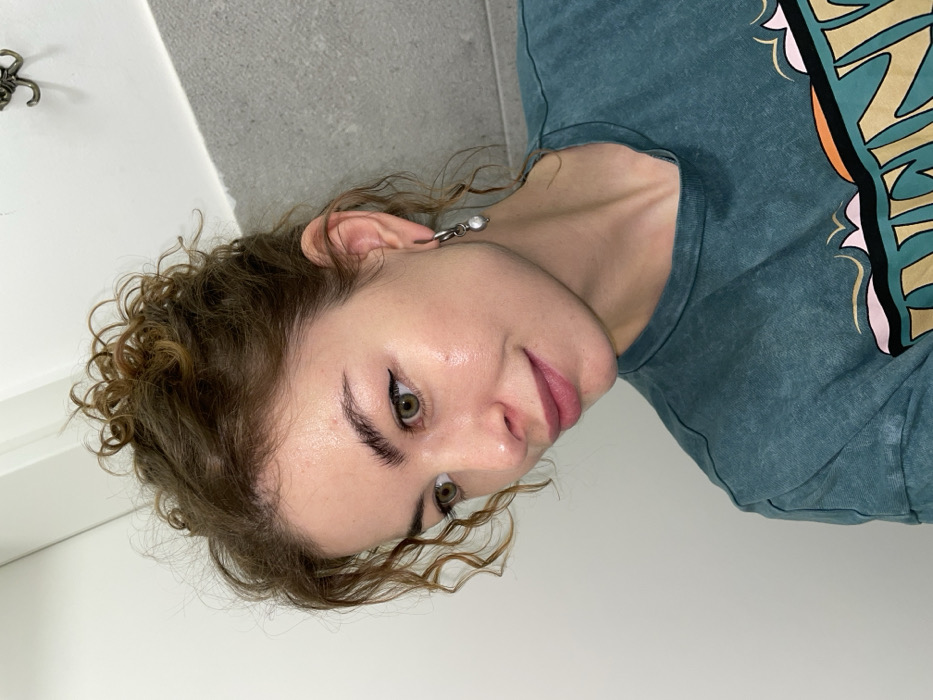 A female individual is depicted in a close-up, chest-up portrait, presumably located in Budva, Montenegro. She is fair-skinned with reddish-brown curly hair styled loosely atop her head, with strands framing her face. Her eyes are light-colored, defined by dark winged eyeliner, and her eyebrows are dark and full. Her lips are a natural pink, and a subtle smile is present. A sheen is visible on her forehead and upper cheeks. She wears a dark teal short-sleeved t-shirt with a partial multi-colored graphic design across the chest, featuring an orange curved element and text. On her visible right ear, a silver-toned earring with a single suspended white pearl is present. The background consists of a plain light-colored vertical surface on the left and a darker gray, possibly concrete, vertical surface on the right. A decorative scrolled metal hook is partially visible in the upper right corner of the gray background.