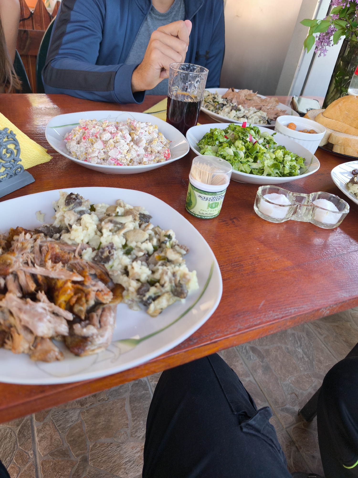 The image depicts a table laden with food, likely in an outdoor setting. Two people are partially visible; one is wearing a blue jacket and the other one is wearing black pants. A variety of dishes are present, including a salad with lettuce and radish, a salad with pink and white ingredients, a dish of shredded meat and mushrooms, and other food items such as bread and sauces. A glass of dark liquid, possibly soda, is on the table, along with a container of toothpicks and a salt shaker. The scene suggests a meal being enjoyed, potentially at a restaurant or cafe. The city and country information provided (Rusokastro, Bulgaria) suggests this is the location of the scene. The table has a reddish-brown wooden surface, and the ground appears to be tiled with brown, stone-like tiles. Lilac flowers are visible in the background.