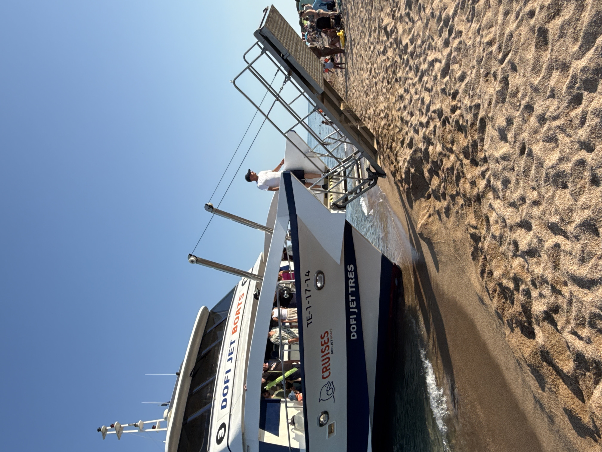 A white and blue catamaran, "DOFI JET TRES," is beached on the sandy shore of Blanes, Spain, with its ramp extended. A person in a white shirt and black cap stands on the ramp, facing the boat. On the boat's deck, several people are visible. The sea laps gently at the boat's hull, and a shadow of the boat is cast on the sand. In the background, more people and beachgoers are scattered on the sand and in the shallow water. The sky is clear and blue.