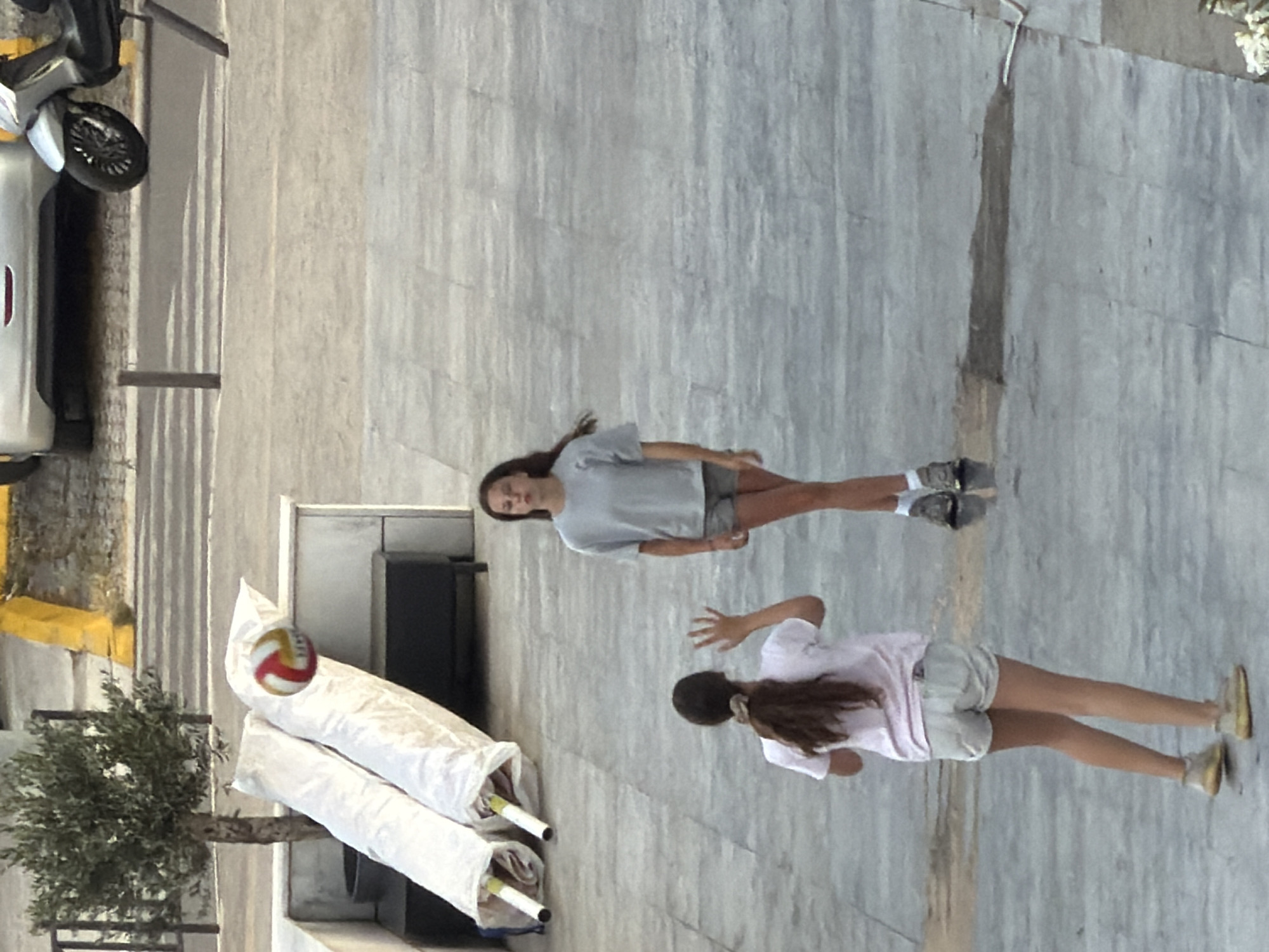 In Budva, Montenegro, two young women are engaged in a game of volleyball on a paved outdoor area. The woman in the foreground, facing away from the camera, wears a white t-shirt and light-colored shorts, her right arm raised as if to receive a hit. Her dark hair is pulled back in a ponytail. The second woman, further back and to the right, is facing the first woman. She wears a gray t-shirt and dark shorts, her arms also in a position to play. A red, white, and yellow volleyball is captured in mid-air above and between the two women. The ground appears to be made of large, light-colored square tiles, with some damp patches visible. In the background, to the left, a small olive tree stands next to two long, white, cylindrical objects, possibly rolled-up mats or canvases. Beyond that, a dark storage unit is partially visible. Further in the background, a silver car and a parked scooter are visible on a street or parking area. The scene appears to be set during daylight, with sunlight casting shadows on the tiled ground.