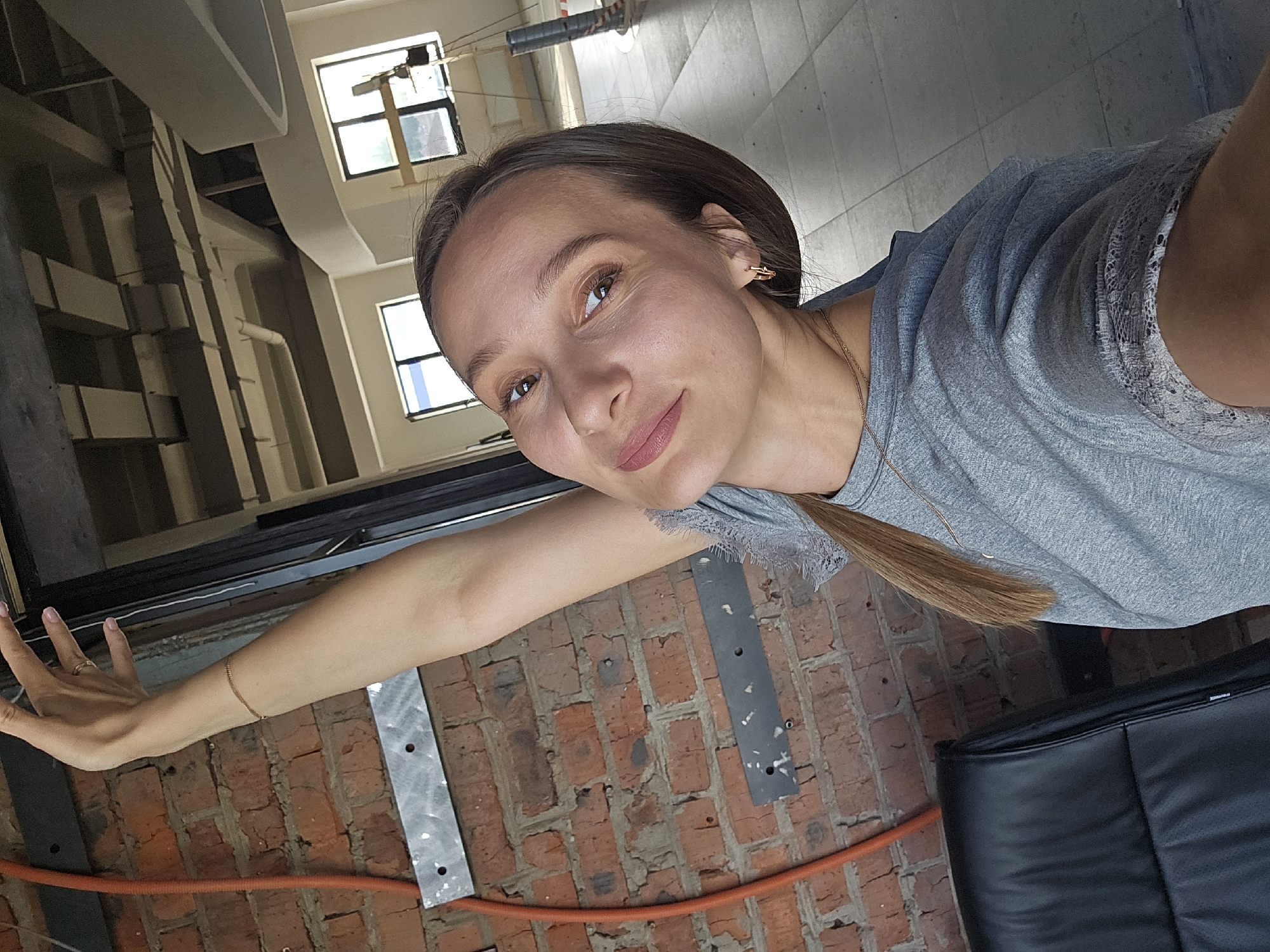 The image is a selfie taken by a young adult woman indoors, captured from a high angle looking down into the room. The woman, with light skin, brown hair, and brown eyes, has a serene expression and a slight smile on her lips, accentuated by light pink lipstick. She wears a grey t-shirt with a delicate lace trim on the sleeve and a thin gold chain necklace. Her left arm is extended towards the left side of the frame, showcasing a thin gold bracelet on her wrist and a ring on her left ring finger; her fingernails are painted a light pink shade.
The background reveals an interior space characterized by an exposed brick wall, suggesting an industrial or loft-style architectural design. High ceilings are visible, with light-colored beams and structural elements, including what appears to be part of a multi-level structure or staircase on the far left. Large windows in the upper portion of the frame allow natural daylight to illuminate the scene, indicating it is daytime. An orange hose or cable lies along the base of the brick wall, and two metallic strips are attached to the wall. A portion of black upholstered furniture is visible in the bottom right corner of the image. The overall atmosphere is casual and bright. The location context provided for this image is Chaikovsky, Russia.
