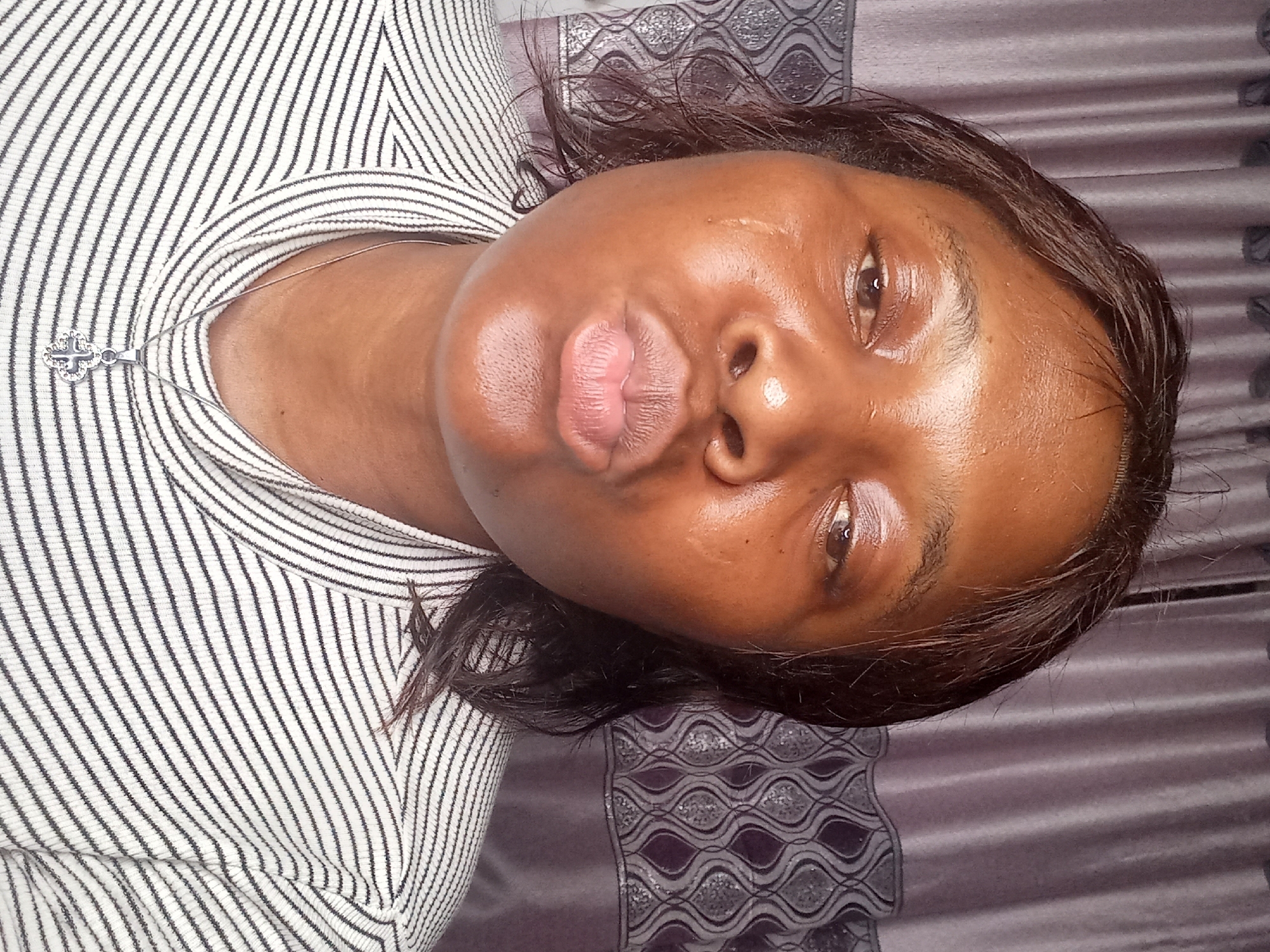 A medium shot shows a woman with dark skin and dark hair leaning forward as if blowing a kiss. She is wearing a black and white striped shirt with a silver necklace with a clover-shaped pendant. The background consists of a dark gray curtain with a lighter gray patterned design.