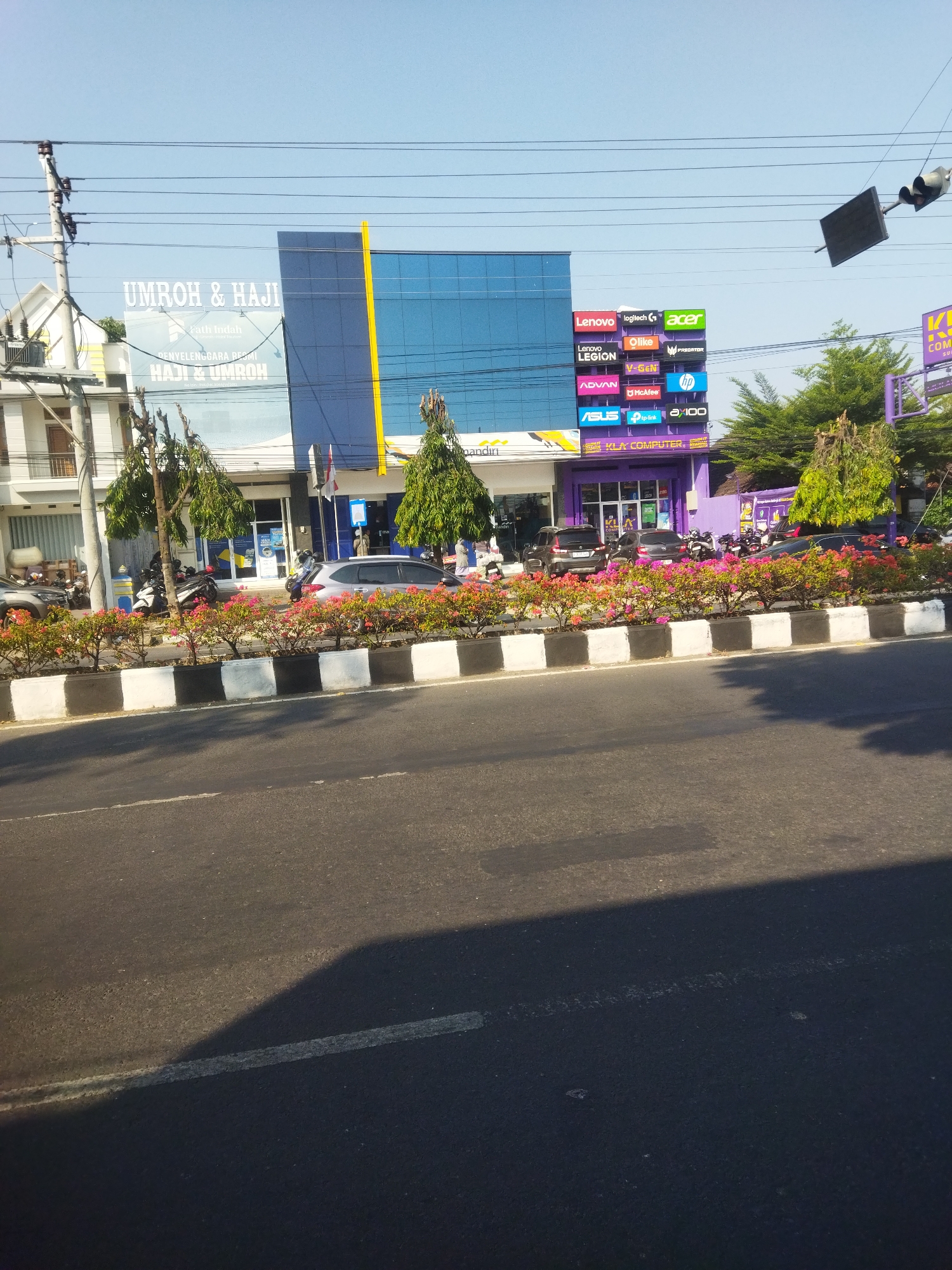 The image depicts a street scene in Sukoharjo, Indonesia, on a sunny day, with a clear blue sky. The focal point is a modern commercial building complex. On the left, a white building with a multi-story facade is visible. Adjacent to it is a prominent blue building with a large vertical yellow accent. A billboard on the blue building displays "UMROH & HAJI" and "Faith Indah Travel & Tourism".
To the right of the blue building, a smaller, vibrant purple building is identifiable as "KLA COMPUTER". Above the purple building, a colorful sign board showcases various technology brands, including Lenovo, Logitech, Acer, Olike, V-Gen, Predator, Advand, McAfee, HP, Asus, TP-Link, and a "a:100" logo.
The street in front of the buildings is lined with a decorative planter filled with pink and red flowers, bordered by black and white painted concrete blocks. Several cars and motorcycles are parked along the roadside and in front of the establishments. People are present, though their identities and actions are not clearly discernible. Traffic lights and power lines are visible in the upper portion of the image, indicating an urban setting. The overall scene suggests a commercial district with various businesses operating.