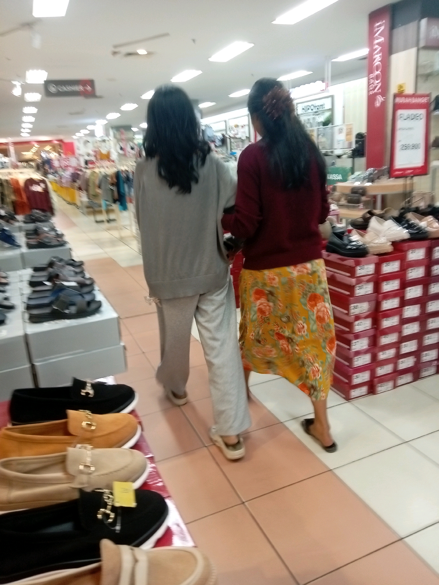 The image shows two women walking in a shoe store. The store appears to be a department store or a mall, with racks of clothing and shelves of shoes visible. The woman on the left is wearing a gray sweater and pants, while the woman on the right is wearing a maroon sweater and a floral print skirt. They are both walking away from the camera, towards the back of the store. In the foreground, there are several pairs of women's shoes on display, including black loafers with gold accents and tan slip-on shoes. The store environment is brightly lit with fluorescent lights. The presence of the "MAROON" branding on a vertical red sign and "HIP-DRIAM" signage suggests this is likely a shoe store within a larger retail complex in Batam, Indonesia, possibly in a shopping mall like Mega Mall or Batam Centre. The overall scene depicts a typical retail environment.