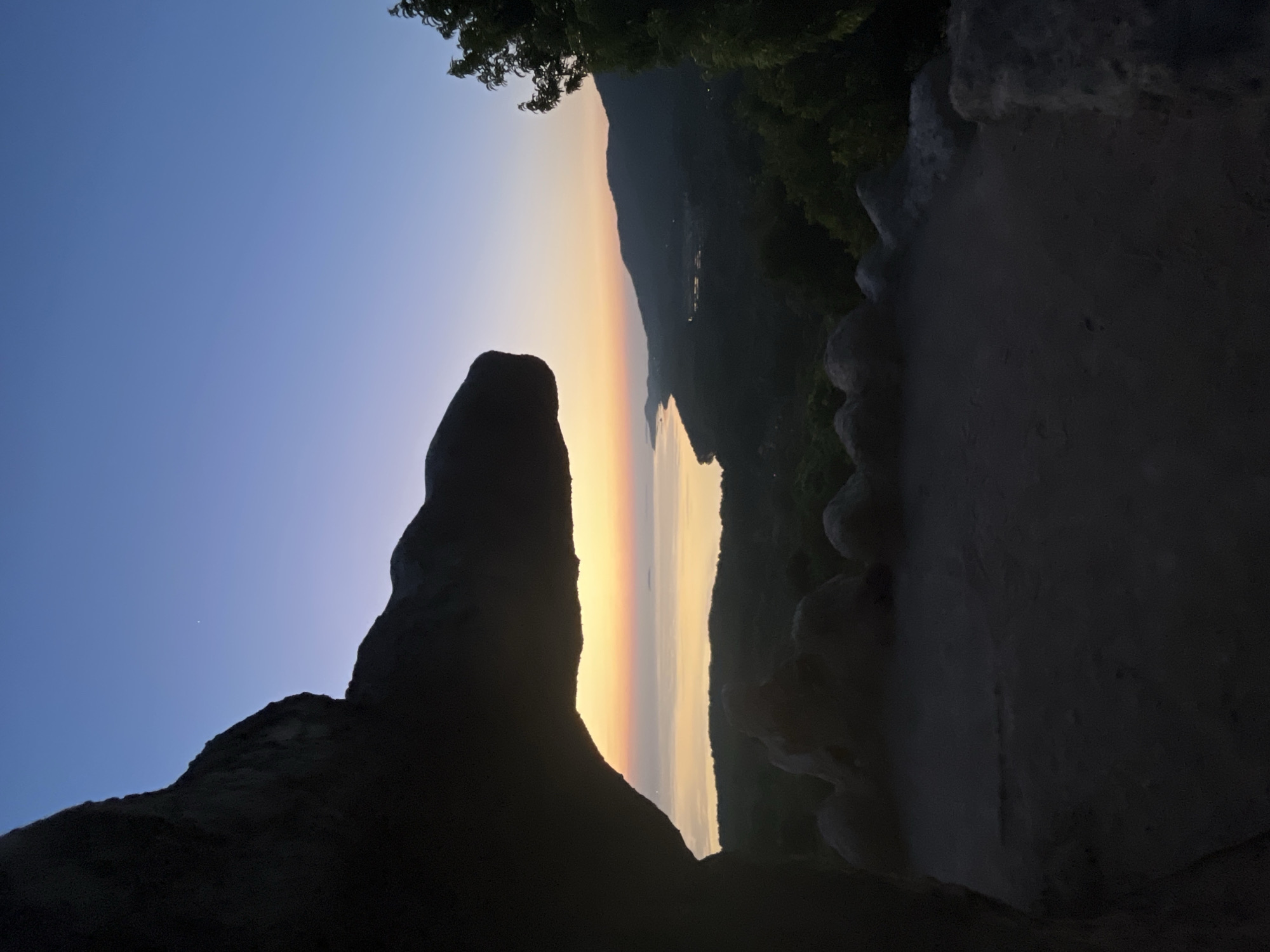 A silhouetted rock formation, resembling a nose and snout, juts into the frame from the left, obscuring a portion of a serene seascape at dawn. The sky transitions from a deep blue at the top to a soft yellow and orange glow at the horizon, where the sea meets a coastline of rolling hills. The water is calm, reflecting the colors of the sunrise. A faint, distant island is visible on the horizon. In the foreground, a rough, textured ground surface is visible, bordered by a low, rocky edge. Lush green foliage is sparsely scattered across the hills and a tree is visible on the far right of the frame. The scene appears to be an overlook, possibly in Karimunjawa, Indonesia, offering a panoramic view of the archipelago at the start of a new day.