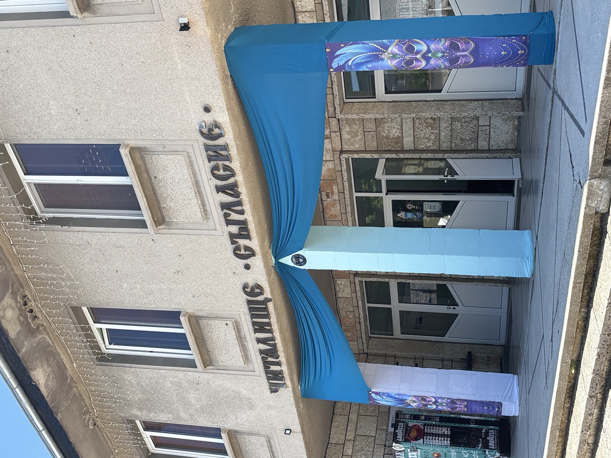 The image shows the entrance to a building with a sign in Bulgarian that reads "ЧИТАЛИЩЕ СЪГЛАСИЕ". This translates to "Chitalishte Saglasie", which is a community center or cultural club. The entrance is decorated with draped blue fabric and two tall pillars covered in light blue fabric, with banners on either side featuring purple masks. To the left of the entrance, a vending machine is visible. The building appears to be made of concrete and stone, with several windows on the upper floor. The overall scene suggests a festive or celebratory atmosphere, possibly related to a carnival or masquerade. The location is identified as the Chitalishte Saglasie in Kavarna, Bulgaria.