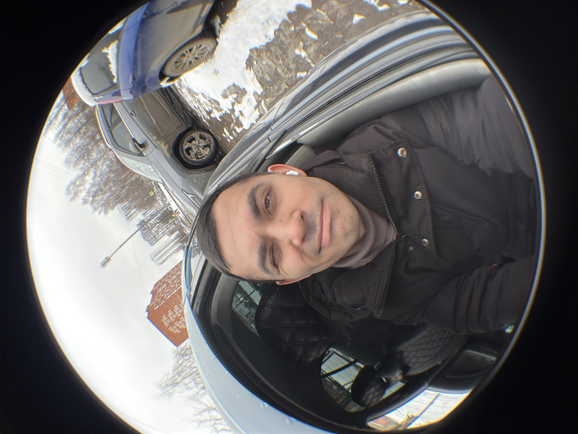 A distorted, wide-angle, selfie-style shot depicts a man sitting in a car, with a view of the external environment suggesting a city location. The man, positioned inside the vehicle, is wearing a dark jacket over a turtleneck sweater and has a wireless earpiece visible in his ear. Outside the car window, other vehicles are parked, with signs of snow or slush on the ground. A brick building and street lighting are visible in the background, adding context to an urban setting. The city of Izhevsk, Russia, is identified as the specific location.