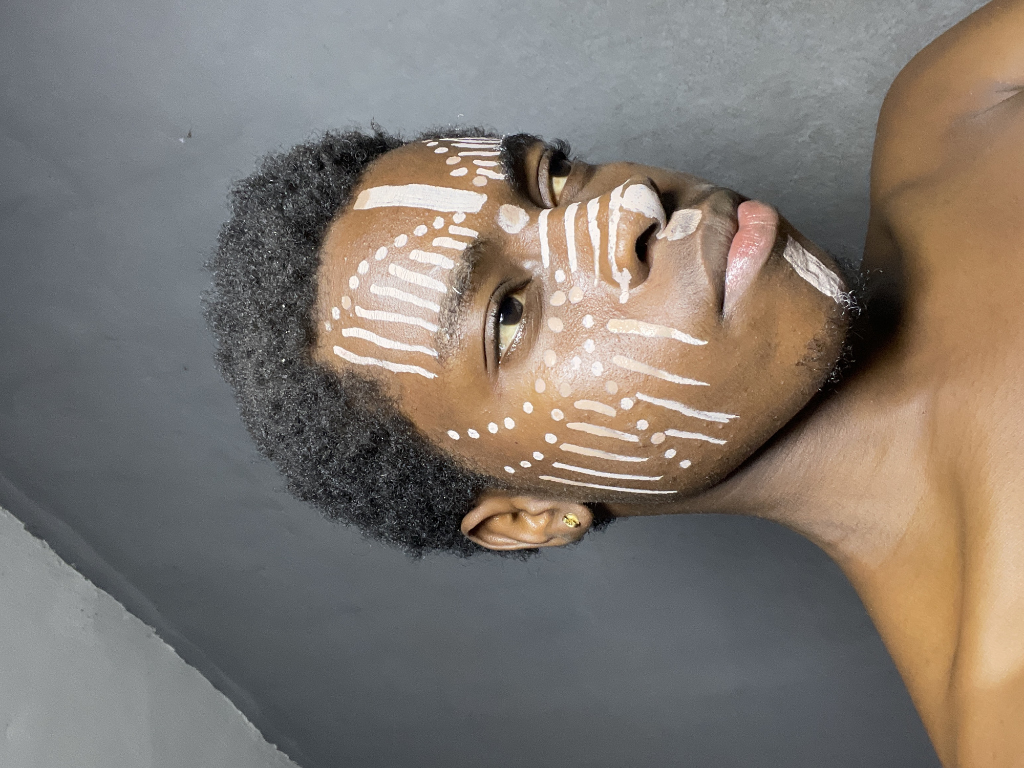A medium shot depicts a young man with dark skin and short, tightly curled black hair, seemingly lying on his back with his head turned toward the camera. His face is adorned with white markings in the form of horizontal lines and dots. He has dark eyes and full lips. A gold earring is visible in his left ear. The background is a muted gray. The location in the city of Nkpor, Nigeria is unknown as it is not visually represented in the image.