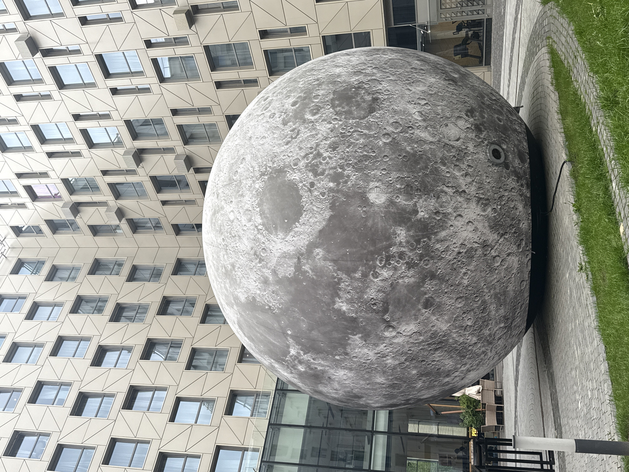 The image depicts a large, spherical sculpture resembling the moon in an urban setting. The sculpture has a gray, textured surface, mimicking the moon's craters and topography. It sits on a paved area, adjacent to a strip of green grass. In the background, there is a modern building with a patterned facade composed of numerous windows. Further back, transparent glass panels can be observed and a possible outdoor restaurant setup with chairs and tables. The photograph shows the object positioned in front of the building, situated on a paved area alongside a strip of grass. The location is Saint Petersburg, Russia.