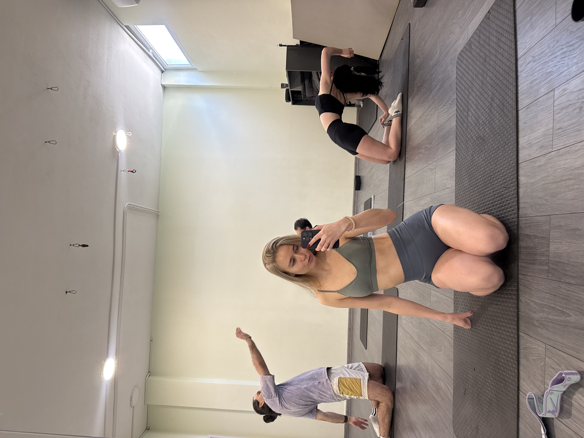 The image shows the interior of a yoga studio, with three individuals engaged in yoga poses and one person taking a selfie with their phone.
The person taking the selfie is in the foreground, kneeling on a yoga mat and wearing a green sports bra and grey shorts. To her left, a man is in a similar kneeling position, doing a yoga pose, wearing a light purple shirt and white shorts with a yellow accent. In the background, further back, is another woman in a backbend pose, wearing a black sports bra and shorts with white socks. All individuals are on separate yoga mats, and the mats are placed on a grey wooden floor that simulates tiles. A locker or storage unit is visible in the background.
The room is brightly lit, with natural light coming through a skylight and artificial light from fixtures on the ceiling. The walls are light beige. There are metal hooks and a lighting fixture on the left wall.