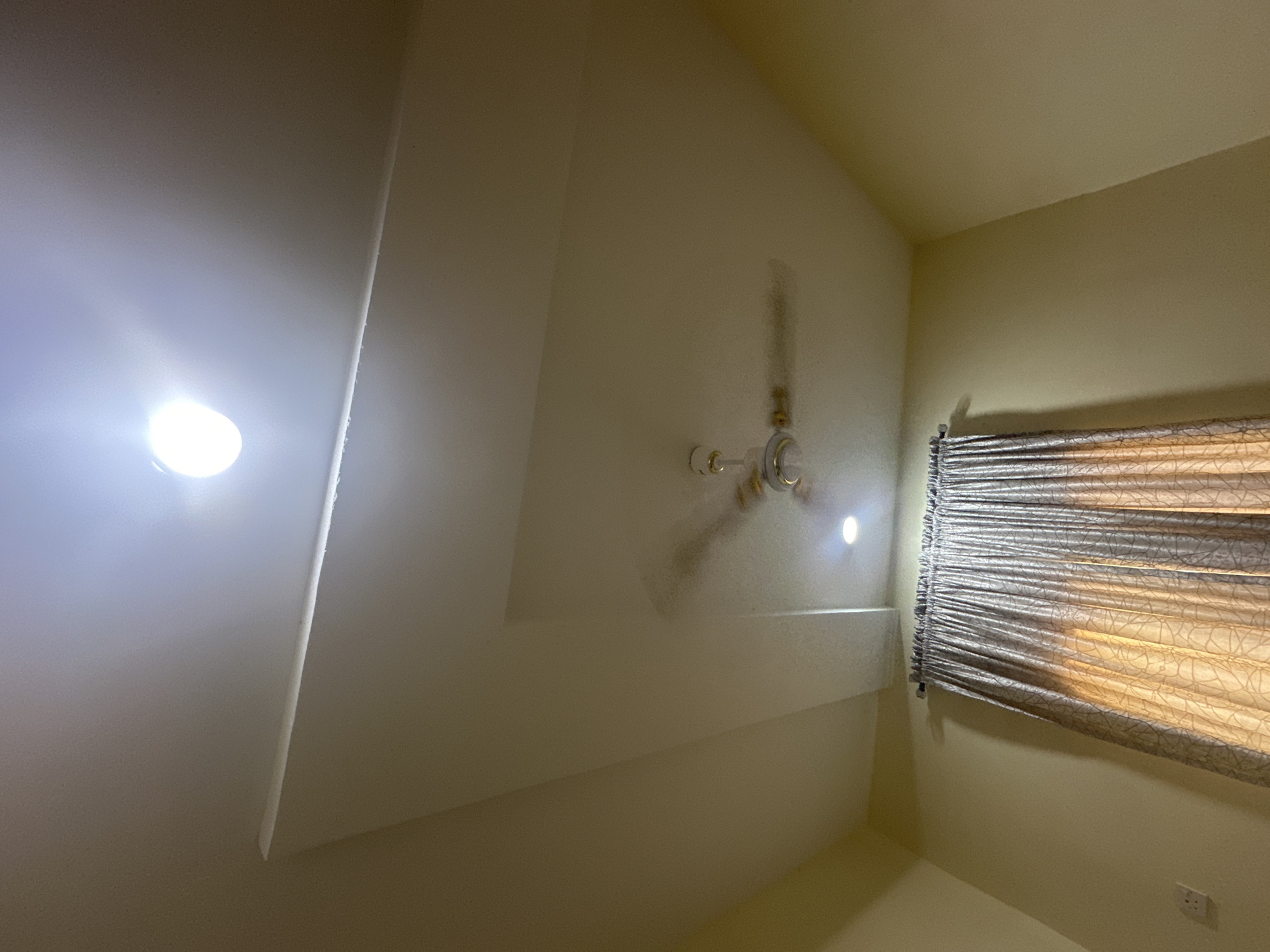 The image depicts the interior of a room, looking upwards towards the ceiling. A recessed ceiling design is visible, with a primary light fixture at the top and a secondary light fixture on the recessed portion. A ceiling fan is centrally located on the recessed ceiling, with its blades blurred due to motion. On the wall below the ceiling, a section of patterned curtains is visible, drawn partially across a window. The walls are painted a light, neutral color. An electrical outlet is visible on the lower left wall. The image appears to be taken in Jalingo, Nigeria.