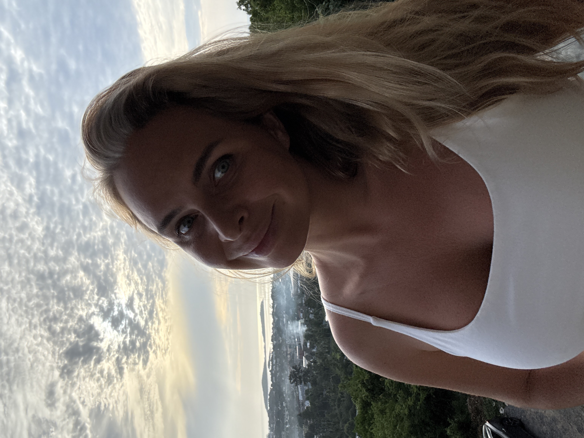 The image shows a woman with blonde hair and blue eyes taking a selfie outdoors. She is wearing a white tank top and smiling at the camera. The background features a scenic view of a tropical landscape with lush green trees, buildings, and the ocean in the distance. The sky is partly cloudy, with the sun casting a warm glow over the scene. The woman appears to be on a balcony or elevated viewpoint, possibly in the city of Ko Samui District, Thailand, given the tropical vegetation and coastal elements.