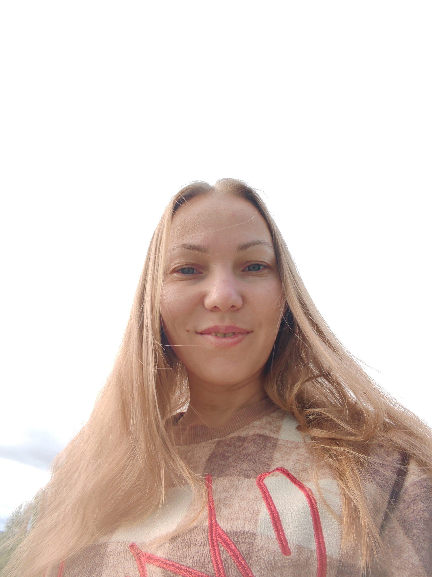 A low-angle, close-up, outdoor photograph shows a young Caucasian woman with long, blonde hair and fair skin, taken from the chest up against a bright, overcast sky. She is looking directly at the camera with a faint smile, her eyes are blue-green, and her lips are slightly parted. A mole is visible on her right cheek. She is wearing a patterned, fuzzy sweater with a plaid design in shades of brown and cream, with bold red embroidery on the front. The background is dominated by a pale, diffused sky, with faint hints of clouds and distant greenery visible at the bottom edges of the frame. The photographer's location is not determinable from the image itself.