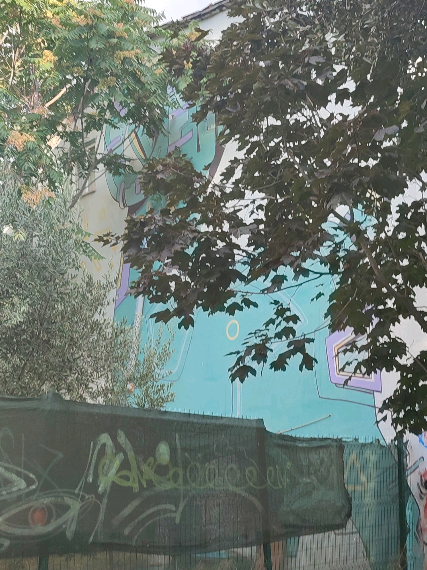 The image depicts a section of a building's exterior wall covered in a large, colorful mural. The mural features abstract shapes and lines in shades of teal, purple, and yellow. Trees with green and dark foliage partially obscure the mural. In the foreground, a dark green mesh fence runs horizontally, also covered in graffiti. The graffiti on the fence includes stylized lettering and an eye-like symbol with a red iris. The background shows parts of the building's facade and the sky. The scene appears to be an outdoor urban environment, possibly in Kadıköy, Turkey, given the context. No people are visible in the image.