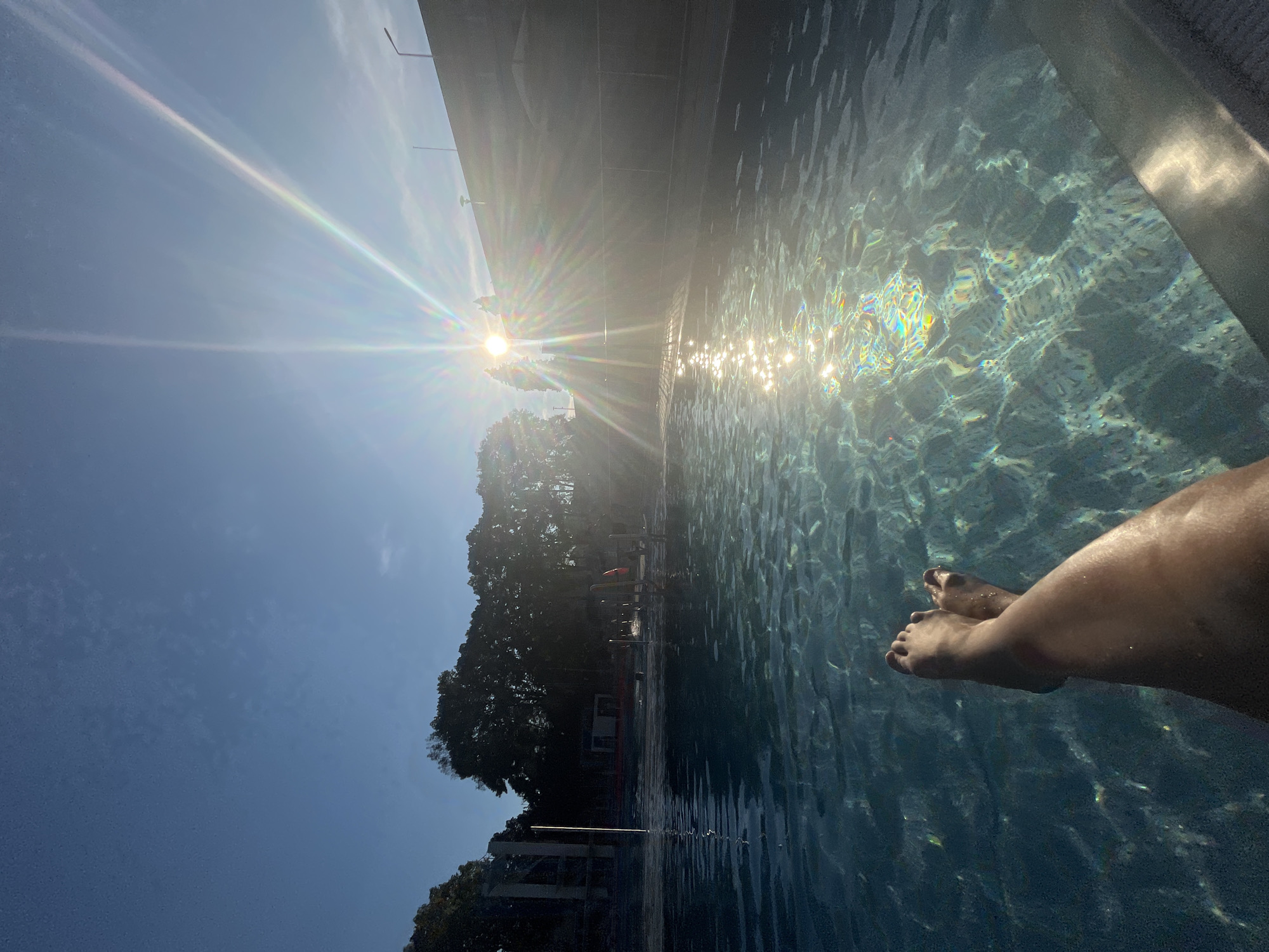 A person's legs and feet are visible in the foreground, submerged in a swimming pool. The water is clear with sunlight glinting off the surface. In the background, trees and buildings are silhouetted against a bright blue sky with the sun directly overhead, creating lens flare. The scene appears to be outdoors in a public or private pool area during daylight.