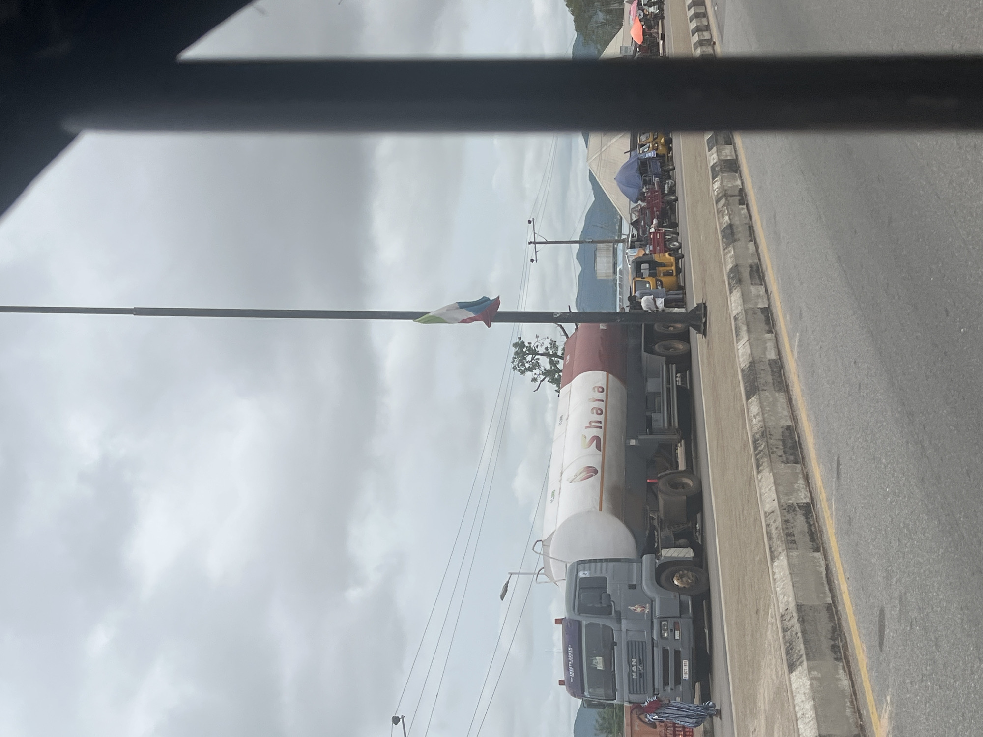 The image depicts a roadside scene in Jalingo, Nigeria. A large grey fuel tanker truck with "Shala" branding is parked on the side of the road. Several people are visible near the truck and on the sidewalk. Behind the tanker, a line of smaller vehicles, including auto-rickshaws (Keke Napep), are also present. In the background, there are tents, trees, and hills under a cloudy sky. A flag is seen on a pole. The overall scene suggests a busy street or marketplace.