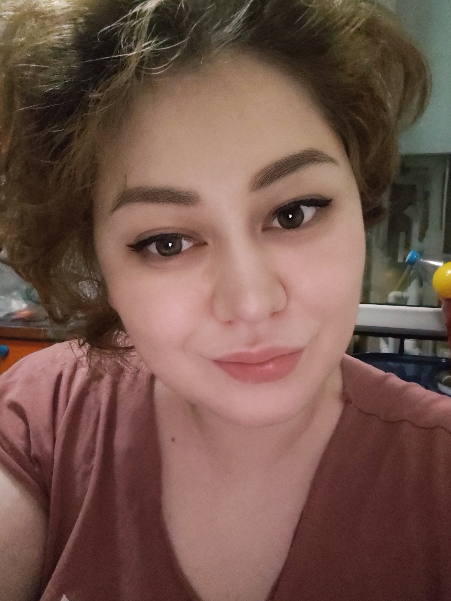 This is a close-up, selfie-style photograph of a woman's face. She has light brown, curly hair, brown eyes, and is wearing makeup, including eyeliner and lipstick. She is wearing a pinkish-brown t-shirt. The background is slightly blurred and appears to be an indoor setting with some objects visible, possibly in a room or kitchen, but the details are indistinct. The photograph does not provide any visual information to determine its location in Fergana City, Uzbekistan.