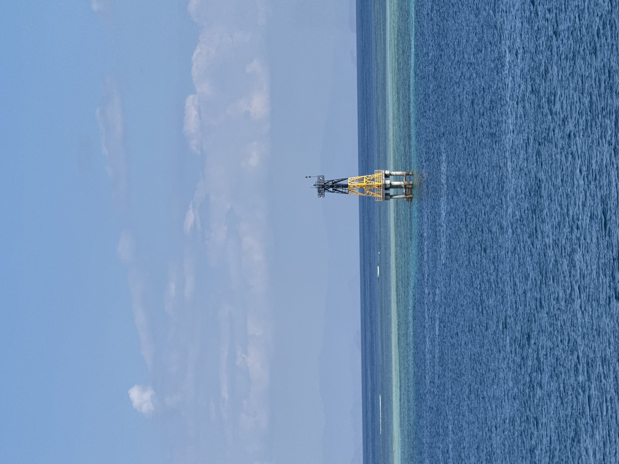 A solitary navigational buoy stands tall in the clear blue ocean off the coast of Gunung Malang, Indonesia. The buoy, a structure of black and yellow metalwork, is supported by four concrete pilings emerging from the water. In the distance, the faint outline of mountains can be seen against a pale blue sky dotted with wispy white clouds. The water surrounding the buoy is a vibrant turquoise, suggesting a shallow coral reef or sandbar beneath the surface. The scene is serene and devoid of human presence, emphasizing the isolation and utility of the buoy in this tranquil seascape.
