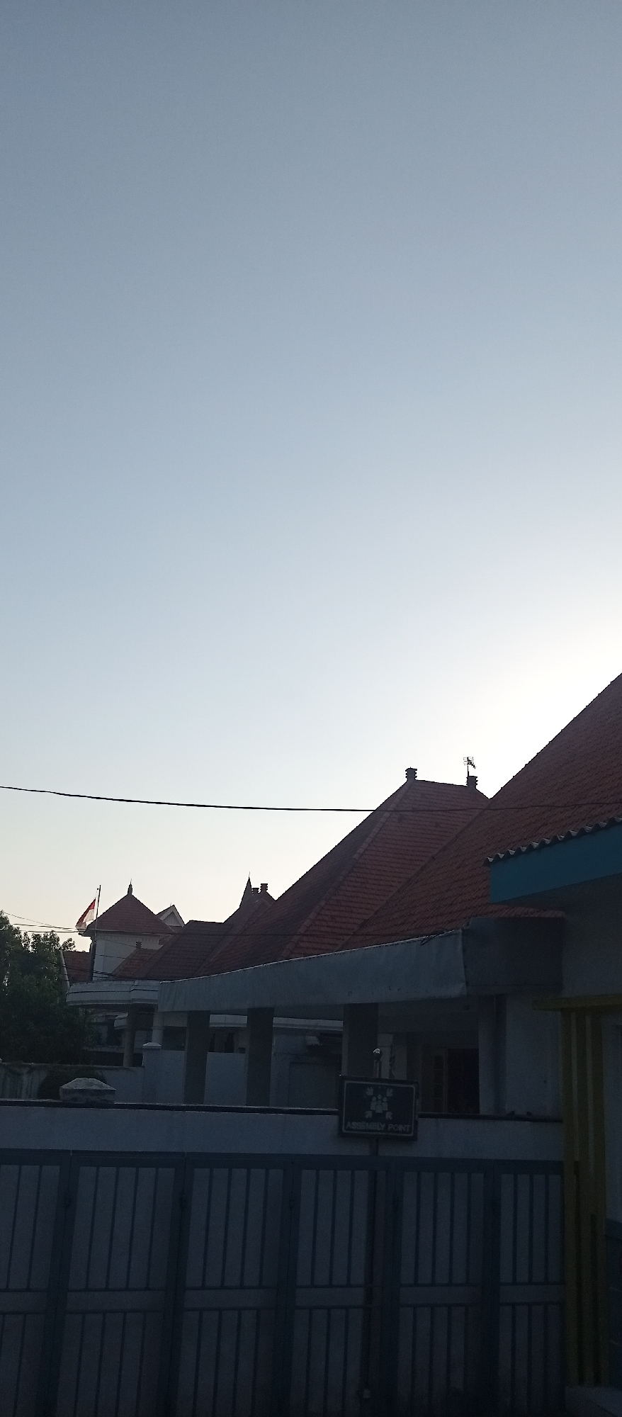 A vertical, medium-shot photograph captures a row of buildings with red-tiled, pitched roofs against a clear, pale blue sky. The sky occupies the majority of the frame, with the buildings positioned in the lower half. A dark, horizontal wire stretches across the sky in the upper middle portion of the image. The architecture appears to be part of a residential or institutional complex, with multiple structures featuring distinct rooflines and protruding eaves. One building on the left has a flagpole with a red and white flag. A sign reading "ASSEMBLY POINT" is visible on a pole in the lower right section, mounted on a wall above a dark metal fence. The overall scene is static, with no discernible human activity. The lighting suggests it is either early morning or late afternoon, with the sun low on the horizon. The location is identified as the Pucangan Village office in Surabaya, Indonesia.