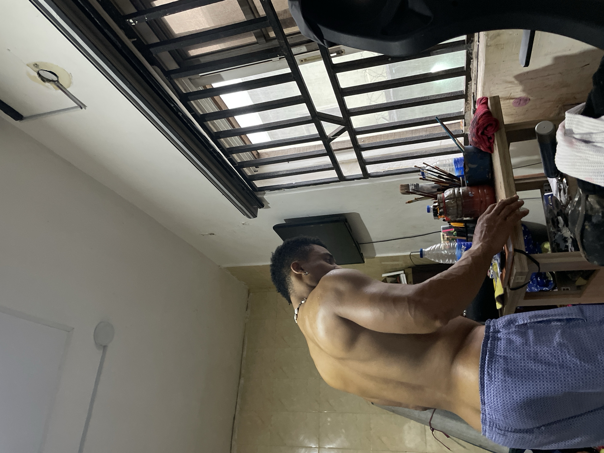 A shirtless man stands in a room with tiled floors and white walls. He wears blue shorts. A metal-framed window with bars is in the upper-center of the image. A small TV hangs from the ceiling. A shelf with art supplies and bottles is beside the man. The location appears to be a room in Ado, Nigeria.