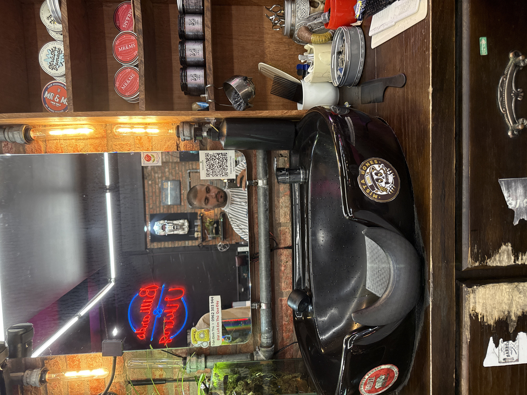 The image shows a barber shop interior. A mirror reflects the interior, showing a man wearing a striped shirt. The reflection also shows a red and blue neon sign "Brouille & Barbers". The mirror is adorned with exposed light bulbs. The reflection includes an advertisement for "Trần Đình Thủ," with the phone number 0962 303 944, and a QR code leading to a booking service. The physical space shows a black barber's sink, grooming products labeled "Mr & Ms", grooming tools, and Reuzel pomade. The name "MISTA BONES" is on a sticker on the black barber's sink. A small "OPEN" sign is visible on a door. The location is identifiable as Đà Nẵng, Vietnam due to the Vietnamese language in the advert and the mention of Trần Đình Thủ.