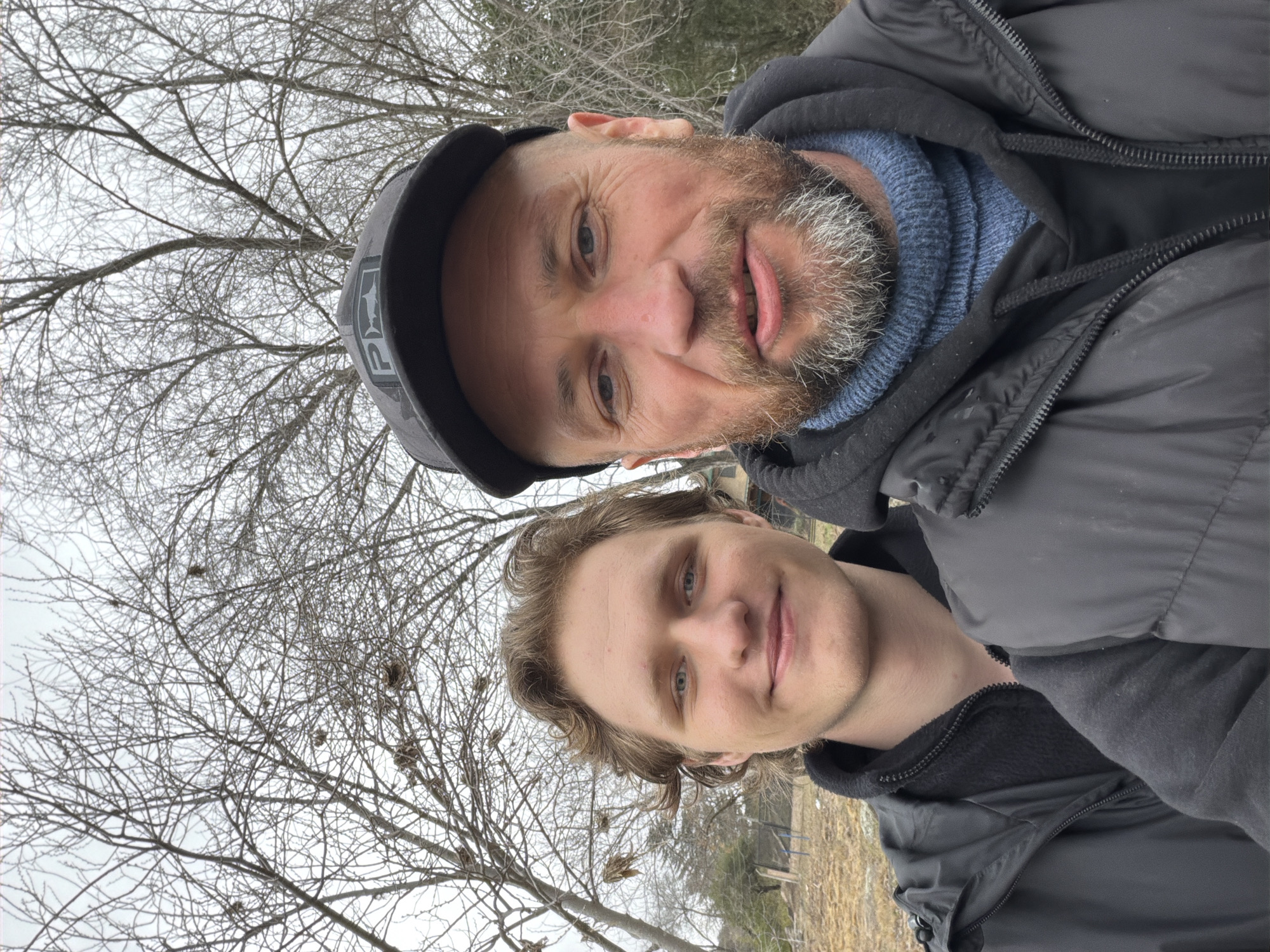 The image depicts a selfie taken outdoors, featuring two individuals. The person in the foreground on the right is a middle-aged man with a beard, wearing a black baseball cap and a dark jacket over a blue sweater. The person on the left is a younger man with wavy hair, smiling and wearing a dark jacket. They are positioned in front of bare trees, suggesting a cool or winter season. The background shows dry grass and some fencing, indicating a rural or semi-rural setting. The overall scene suggests a casual outdoor activity. The image was taken in Comuna Estancia Vieja, Argentina.