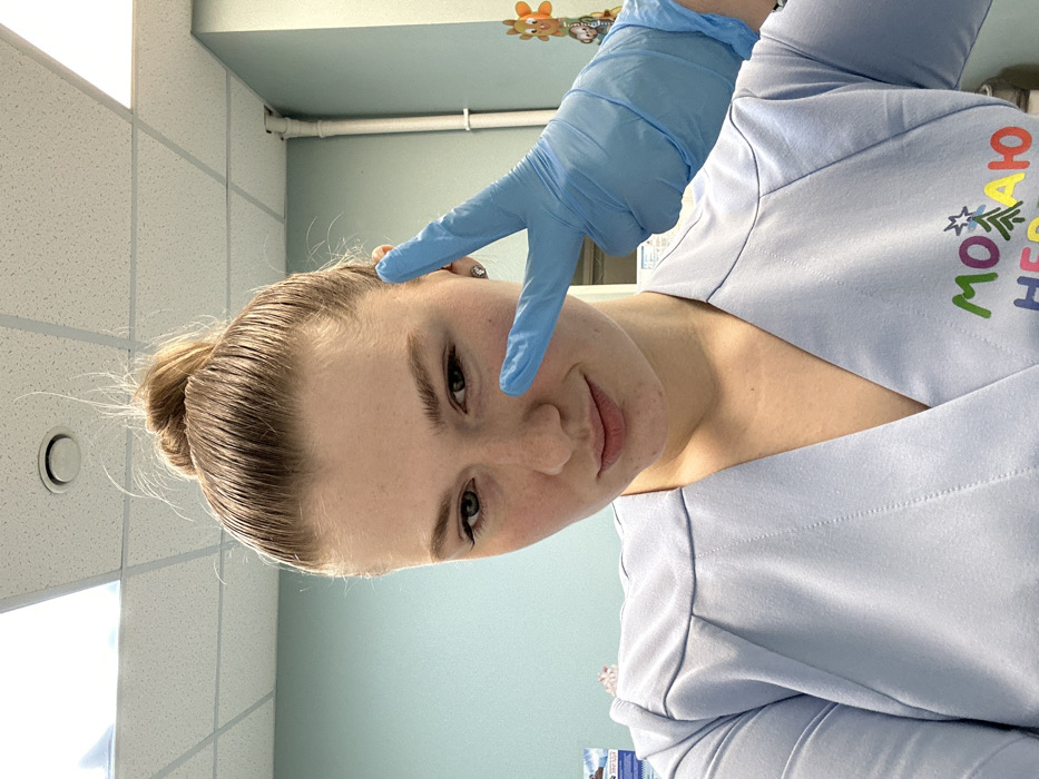 A young woman with blonde hair pulled back in a bun poses for a selfie. She wears light blue scrubs with colorful Cyrillic lettering on the right side. Her left hand, covered in a light blue glove, is held up to her face in a peace sign. The background includes a tiled ceiling and light blue walls. A few decorative items are visible in the upper background. The image is tilted slightly.
