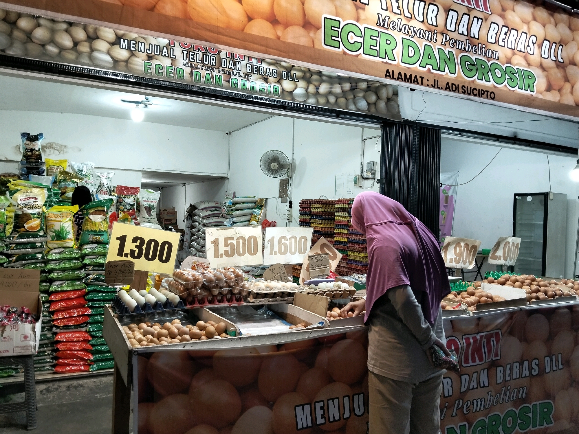 This is a daytime, eye-level, medium shot of a food stall, likely an egg and rice seller, in Arang Limbung, Indonesia. The stall is covered by a large sign that reads "MENJUAL TELUR DAN BERAS DLL" (Selling Eggs and Rice etc.) and "MELAYANI PEMBELIAN ECER DAN GROSIR" (Serving Retail and Wholesale Purchases). The stall appears to be situated along a street, with a closed metallic gate visible on the left.
The foreground is dominated by a counter displaying various types of eggs in plastic trays and loose in wooden crates. A woman wearing a purple hijab and light-colored pants is standing at the counter, partially obscured by the display. She appears to be selecting or inspecting eggs, with her left hand reaching into a crate. Her right hand is tucked into the back of her pants, holding a small, patterned pouch.
Behind the counter, numerous sacks of rice are stacked, some in various colors like green and red. Several yellow signs with prices are visible, including one that reads "1.300" prominently displayed near the eggs. Other signs show prices like "1.500" and "1.600".
In the background, more shelves and displays of goods are visible. A ceiling fan with a light bulb is mounted above the stall. The overall impression is that of a busy, local market setting.