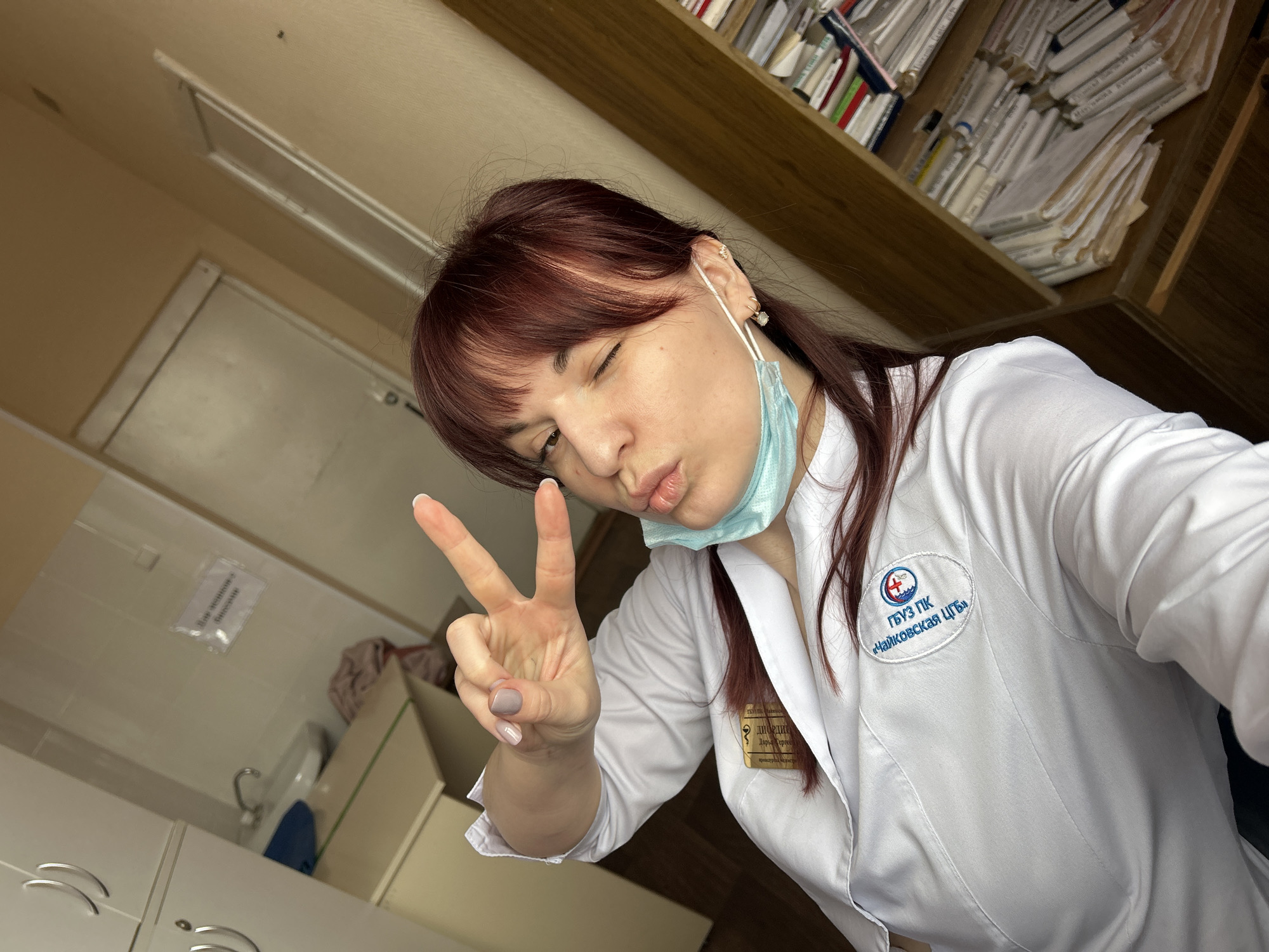 A woman in a white lab coat is taking a selfie in what appears to be a medical office. She is winking, making a peace sign with her left hand, and pursing her lips. She is wearing a light blue medical mask, pulled down below her mouth.
The background includes a shelf filled with files, cabinets, a sink, and a sign in Russian. The logo on the lab coat indicates the location is connected to the city of Чайковский (Chaykovsky). Based on the text on the badge, it also indicates a place named Днородися Дарья Сергеевна, which is likely a place of medical care located in the city of Фоки, Russia.