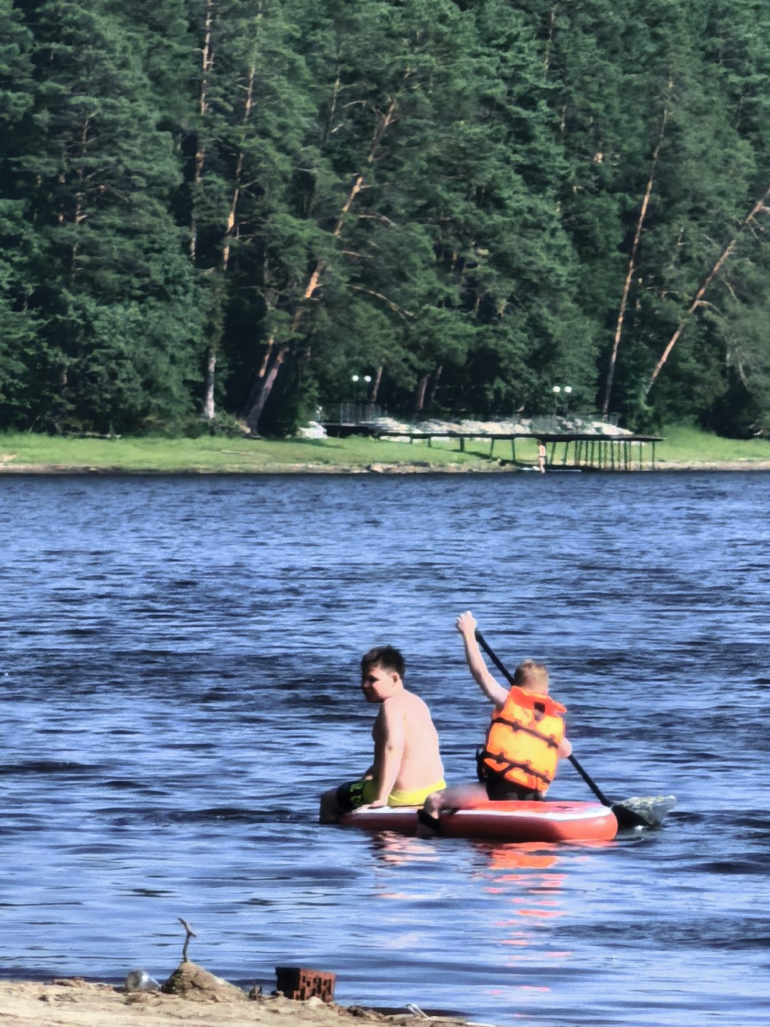 Two young males are paddleboarding on a wide, calm body of water, likely a river or lake, with a dense, green forest lining the far bank. The male in the foreground, on the left, is shirtless and wearing yellow swim trunks. He is seated and facing away from the camera. The male on the right is also shirtless and is wearing an orange life vest. He is seated further back on the paddleboard and is holding a paddle, appearing to be propelling them through the water. The paddleboard is red and white. In the background, the forest is thick with tall trees. Along the shoreline of the forest, there is a wooden structure, possibly a pier or dock, with several upright posts. The water is a deep blue, reflecting the sunlight. The overall scene suggests a warm, sunny day spent outdoors. There is no specific information available that places this scene within the city of Asbest, Russia.