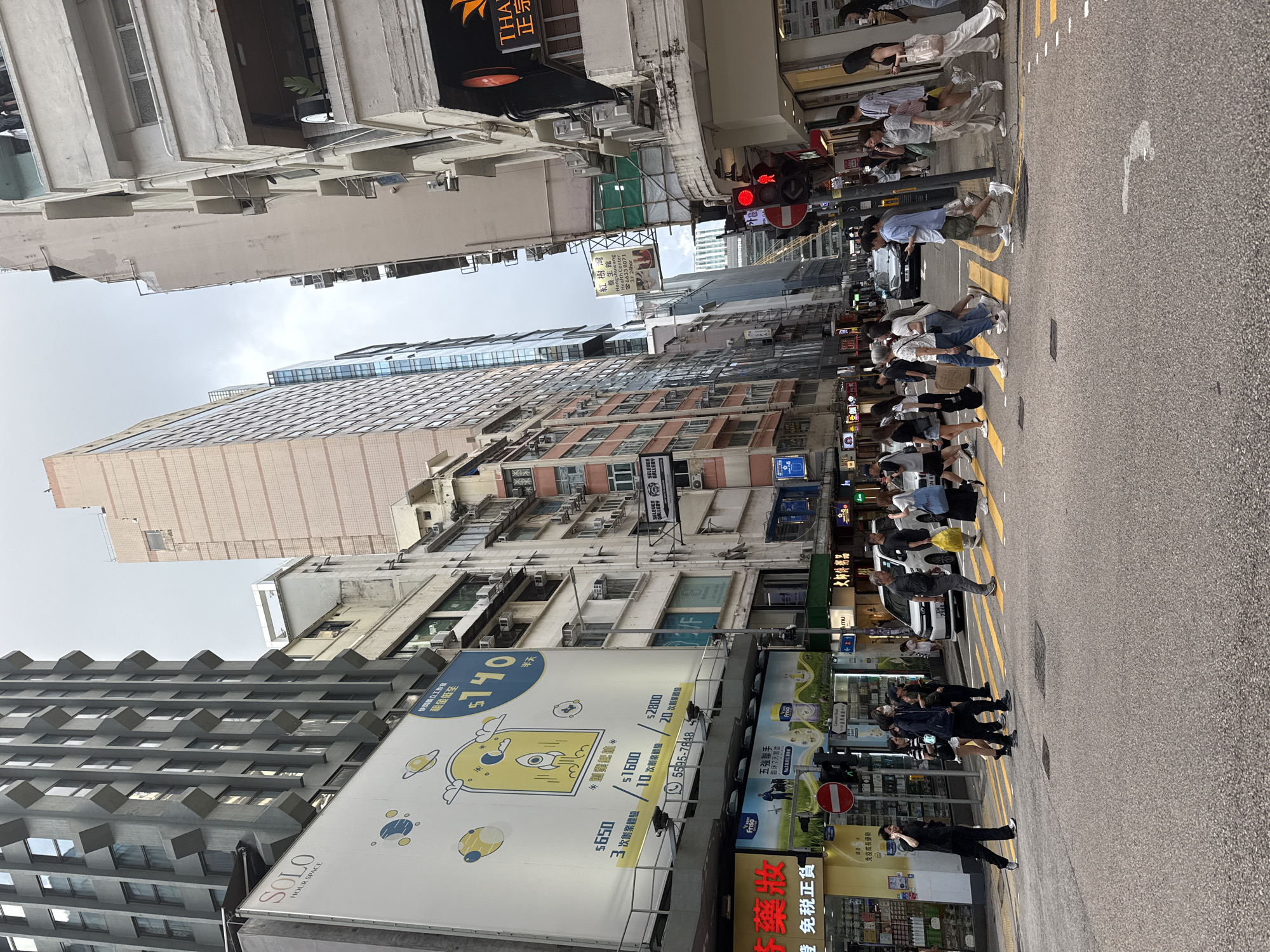 The image depicts a street scene in Hong Kong, China, likely in a commercial district, characterized by tall buildings on either side of a road. The sky is overcast. A pedestrian crosswalk is centrally located, with several people crossing in various directions. On the left side of the street, a large billboard advertises "SOLO HOUR SPACE" with graphics of planets and a rocket. Below this, there are more advertisements for products like "Friso" milk. Traffic lights are visible, with one showing a red pedestrian signal. A white van and a black car are stopped at the intersection. People in the foreground are walking across the crosswalk, some carrying bags. Buildings feature a mix of modern and older architectural styles, with numerous windows and air conditioning units visible. Shops with signage in Chinese characters are present at street level. The overall impression is of a busy urban environment.