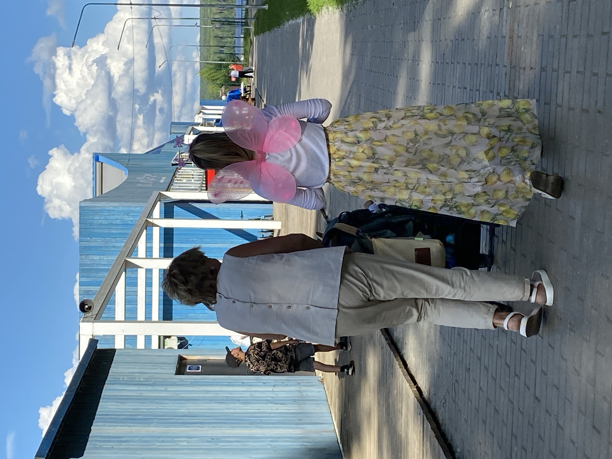 In Chaikovsky, Russia, on a sunny day with cumulus clouds, two women walk away from the camera on a brick pathway. The woman in the foreground, from the back, wears a light-colored sleeveless top with buttons down the back and light-colored trousers, paired with white sandals. She is pushing a stroller. To her right, a younger woman wears a white long-sleeved shirt and a long yellow skirt with a lemon print. She has pink, translucent fairy wings attached to her back. Behind them, to the left, a young boy in a patterned shirt and shorts stands looking to his left. The environment includes a long, low building with blue wooden siding and white trim, and further down the path, more buildings and lampposts are visible, with a body of water and trees in the distance.