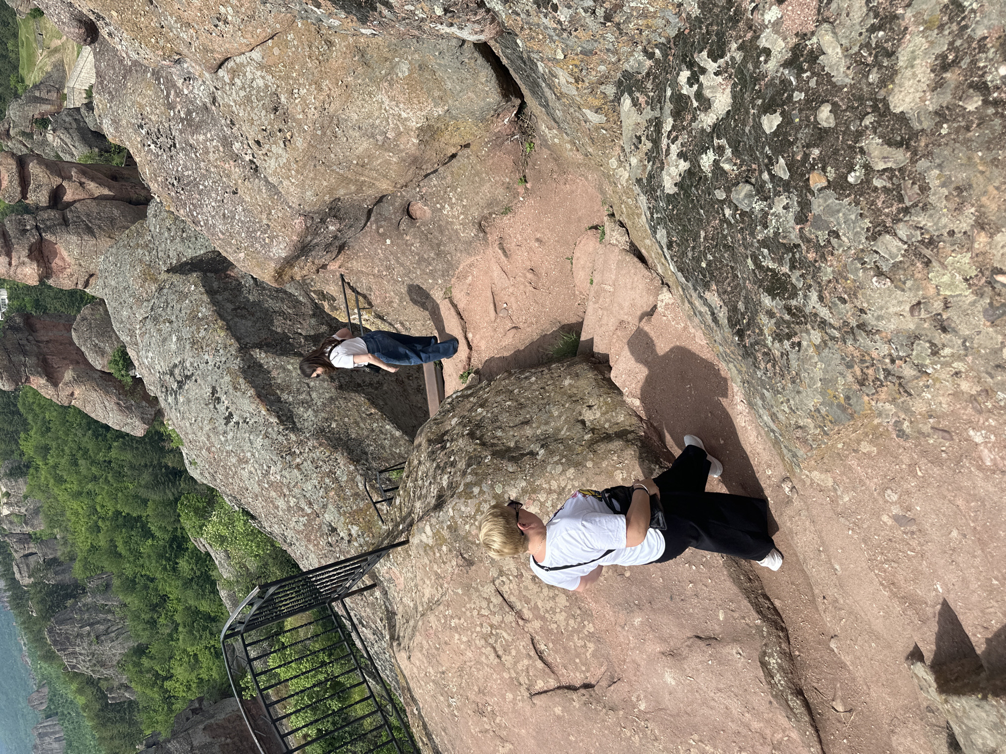 The image shows an elevated outdoor scene in Belogradchik, Bulgaria. Two women are present on a rocky, mountainous terrain. One woman is standing on a metal platform and wears a white shirt and blue jeans. Another woman is standing on a rock surface, wearing a white shirt and black pants. A metal railing borders one side of the rocky area. The landscape includes rocks and green vegetation.